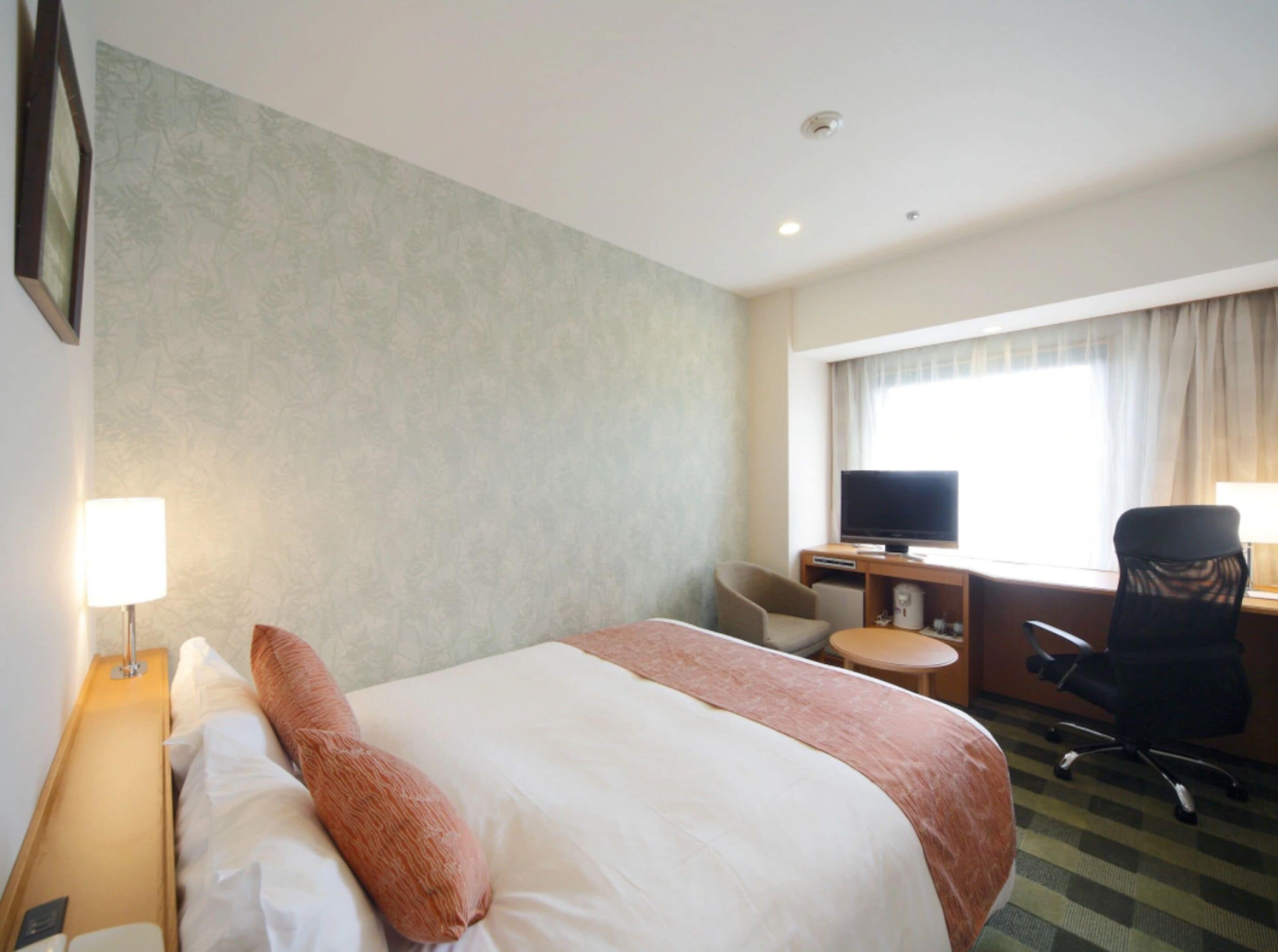 A hotel room with a bed, a desk with a television, a chair, and a window with curtains.