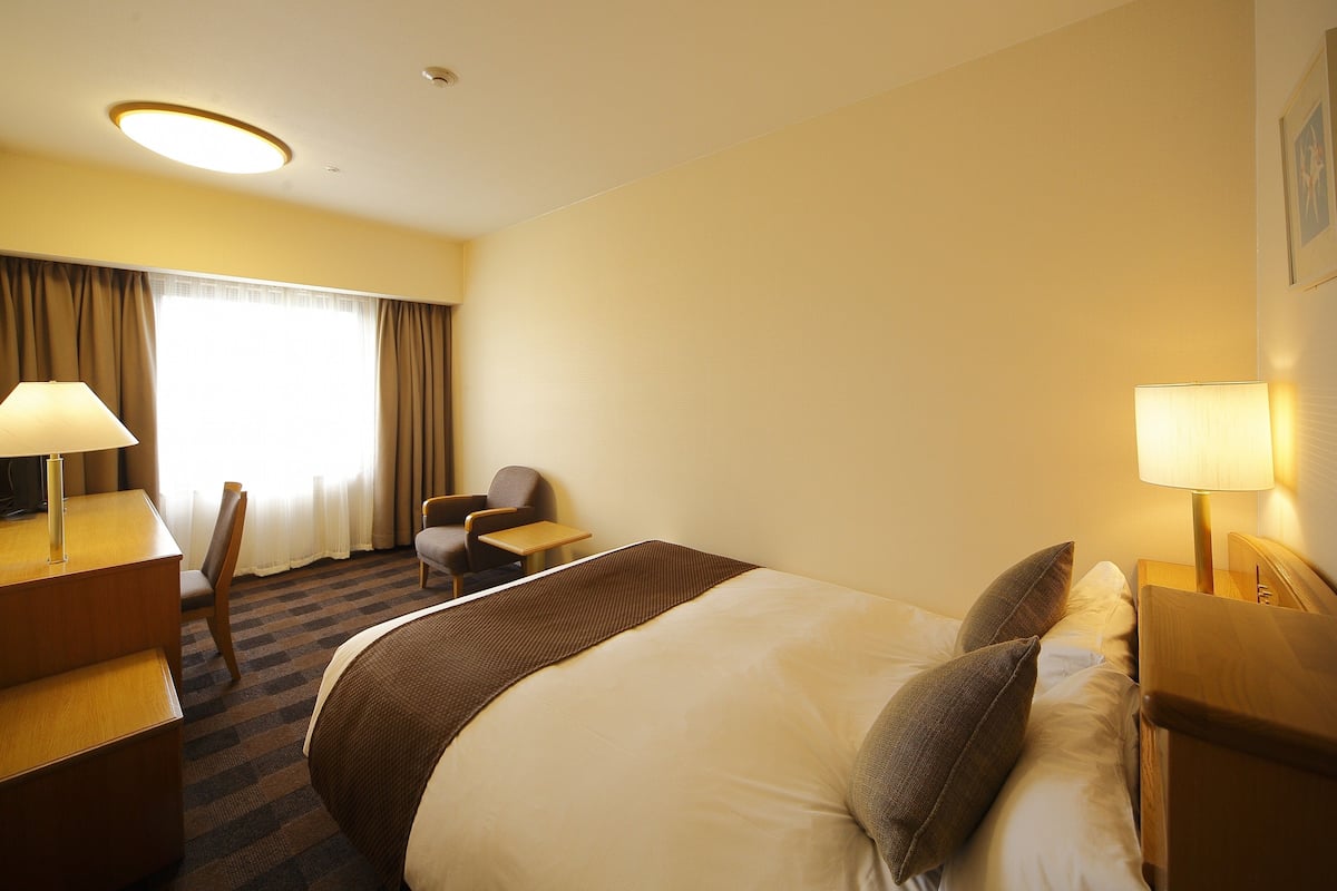 Une chambre d’hôtel comprenant un lit, deux tables de chevet, un bureau avec une chaise, une lampe et une fenêtre avec des rideaux.