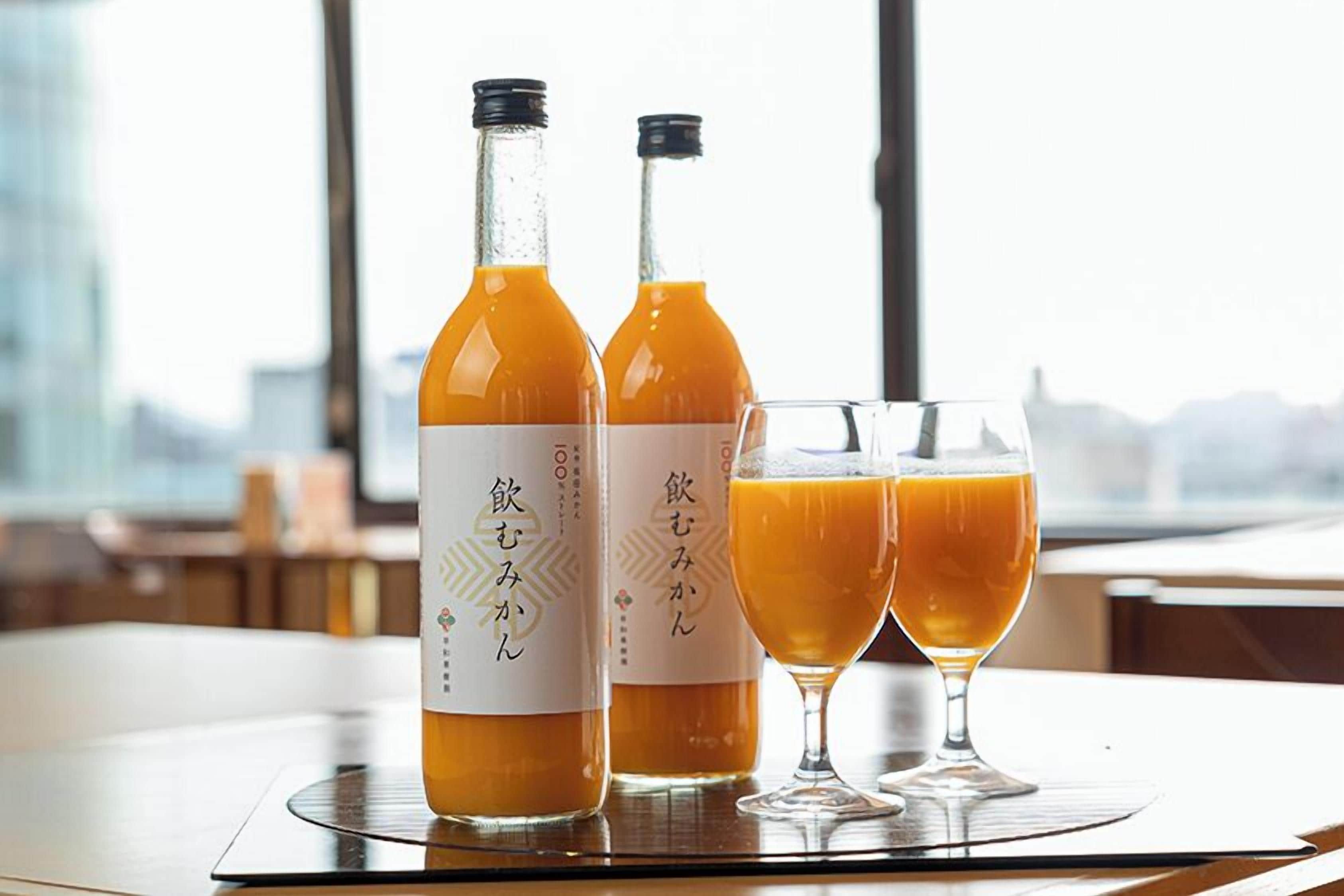 Two bottles of orange juice and two glasses on a table with a view of a cityscape.