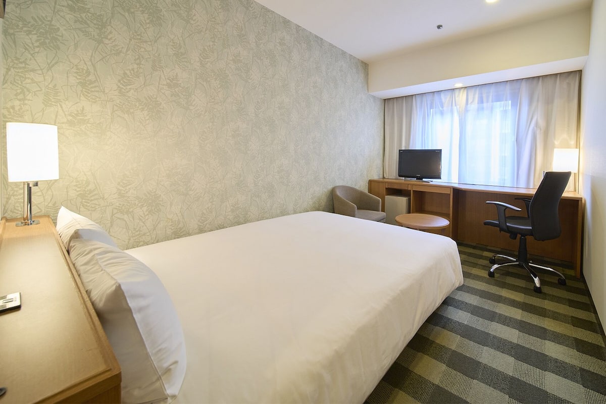 Een hotelkamer met een bed, een bureau met stoel, een televisie en een raam met gordijnen.