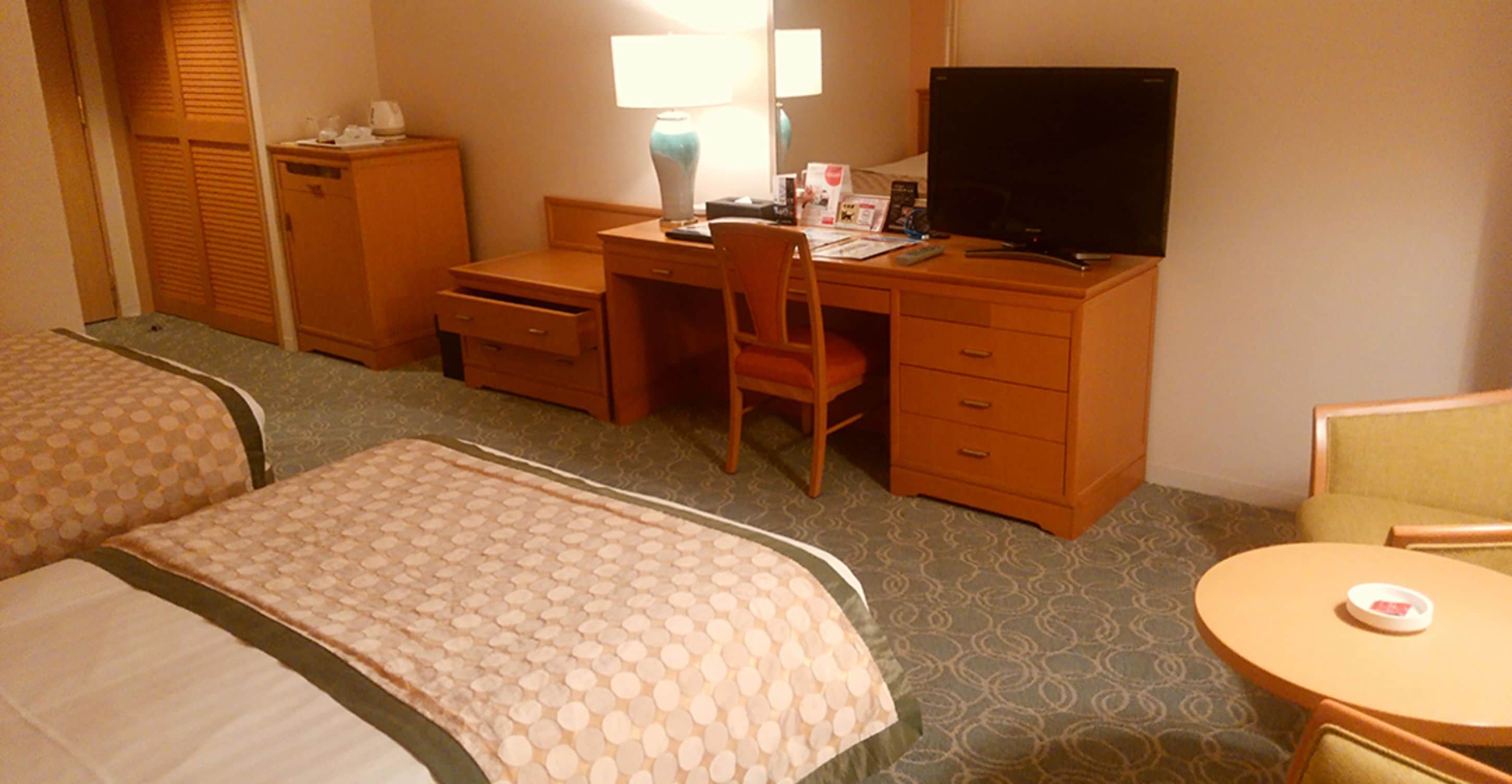 Ein Hotelzimmer mit zwei Betten, einem Schreibtisch mit Stuhl, einem Fernseher und einem kleinen Tisch.