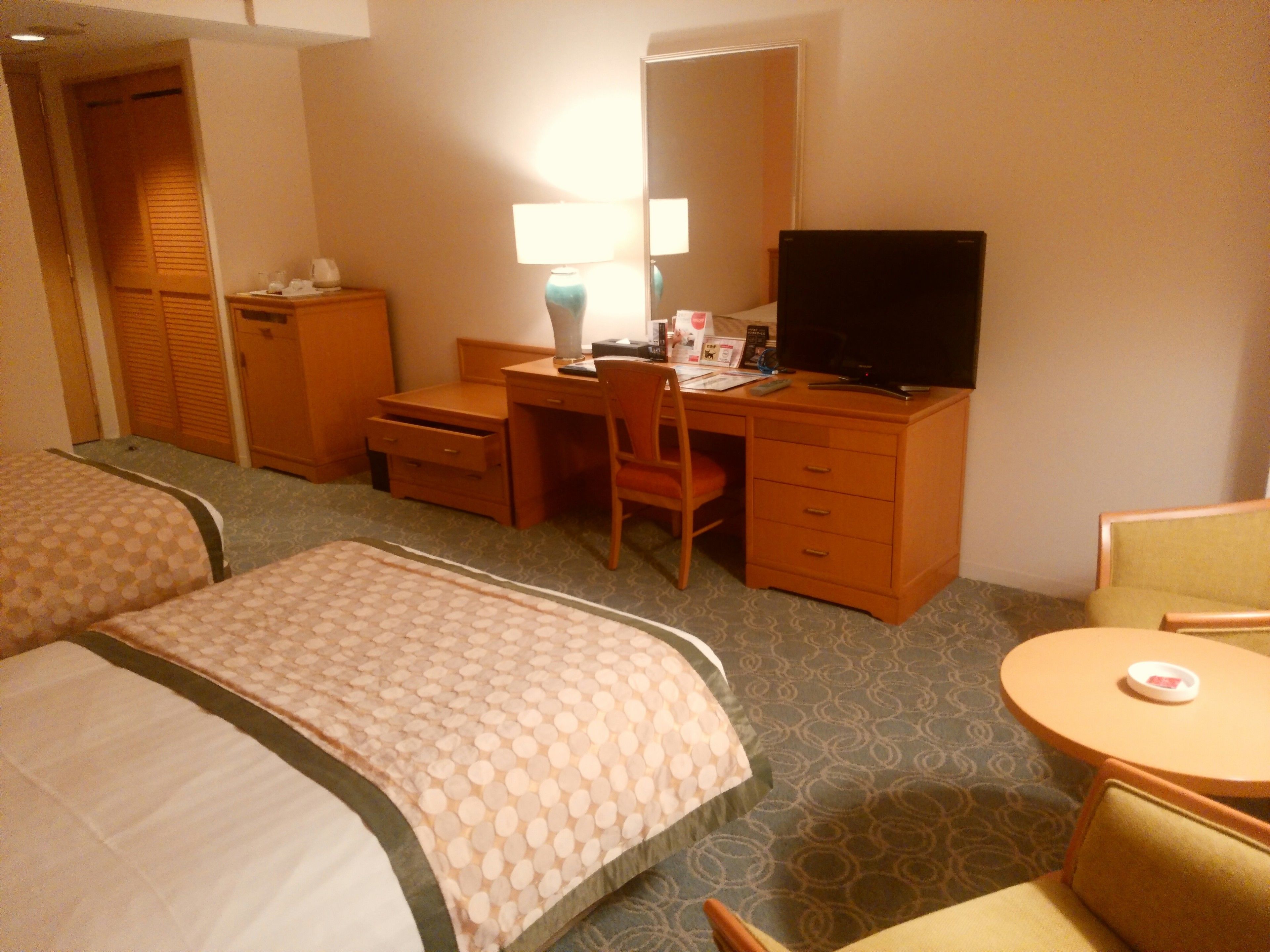 A hotel room with two beds, a desk with a computer, a chair, a TV, and a small round table.