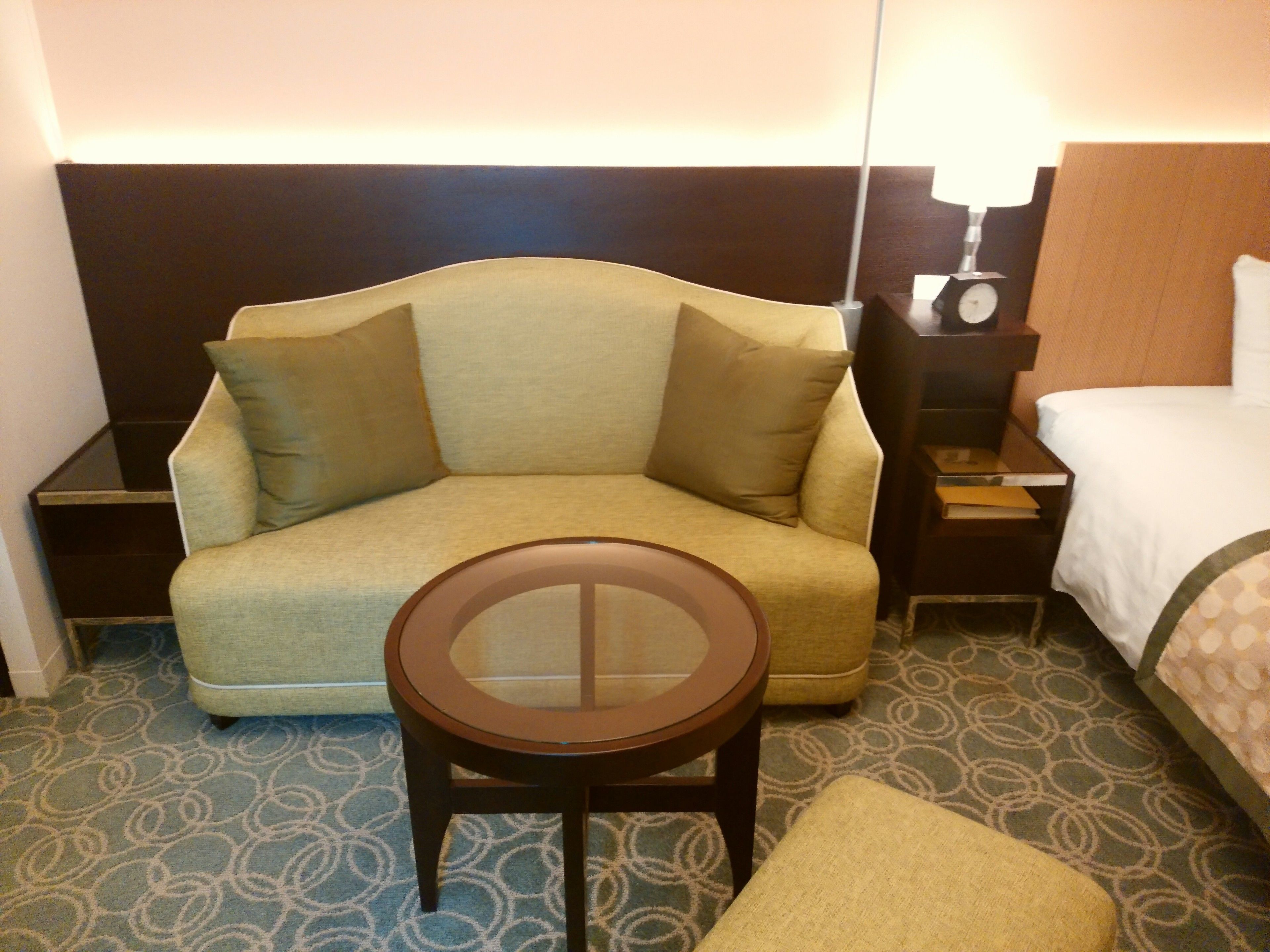 A hotel room with a sofa, a small round table, a bedside table with a lamp, and a bed.