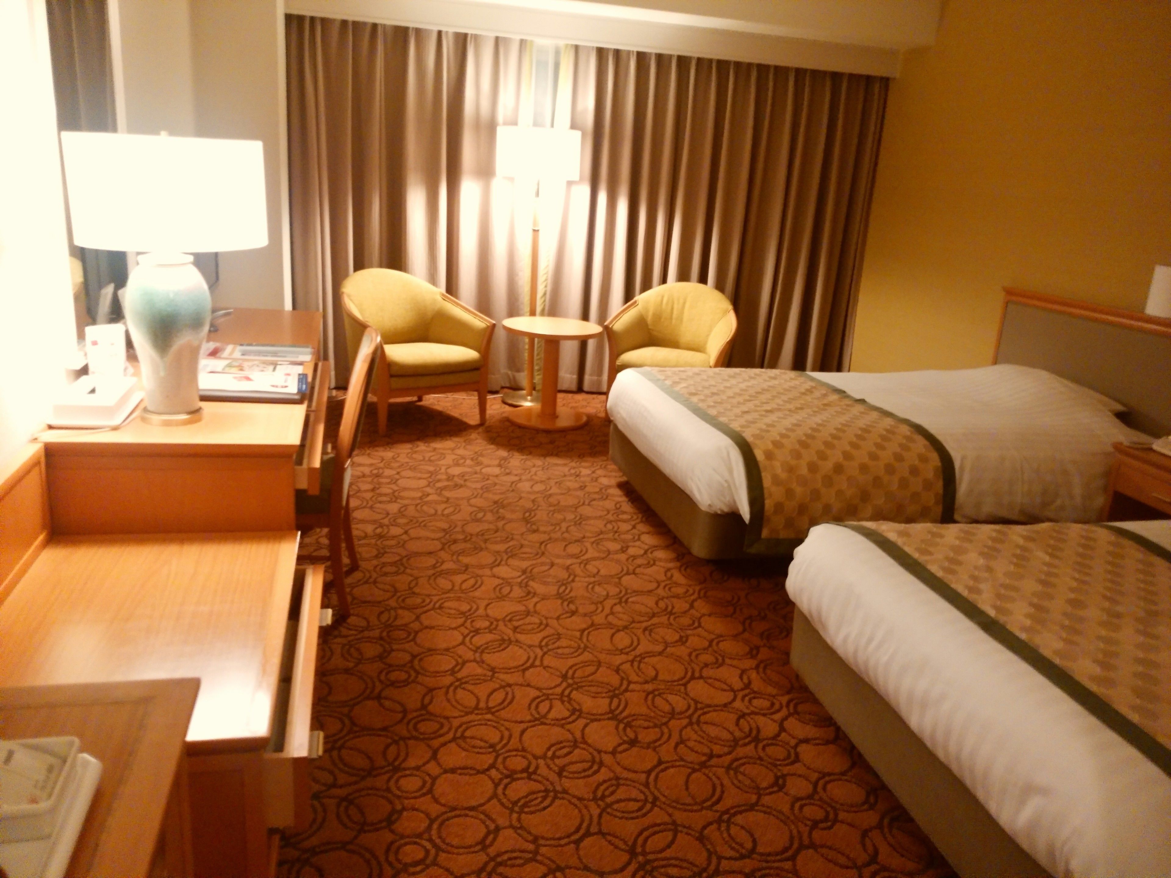 A hotel room with two single beds, a desk with a lamp, two armchairs, and a small round table.