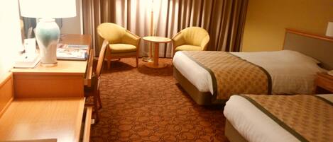 A hotel room with two single beds, a desk with a lamp, two armchairs, and a small round table.