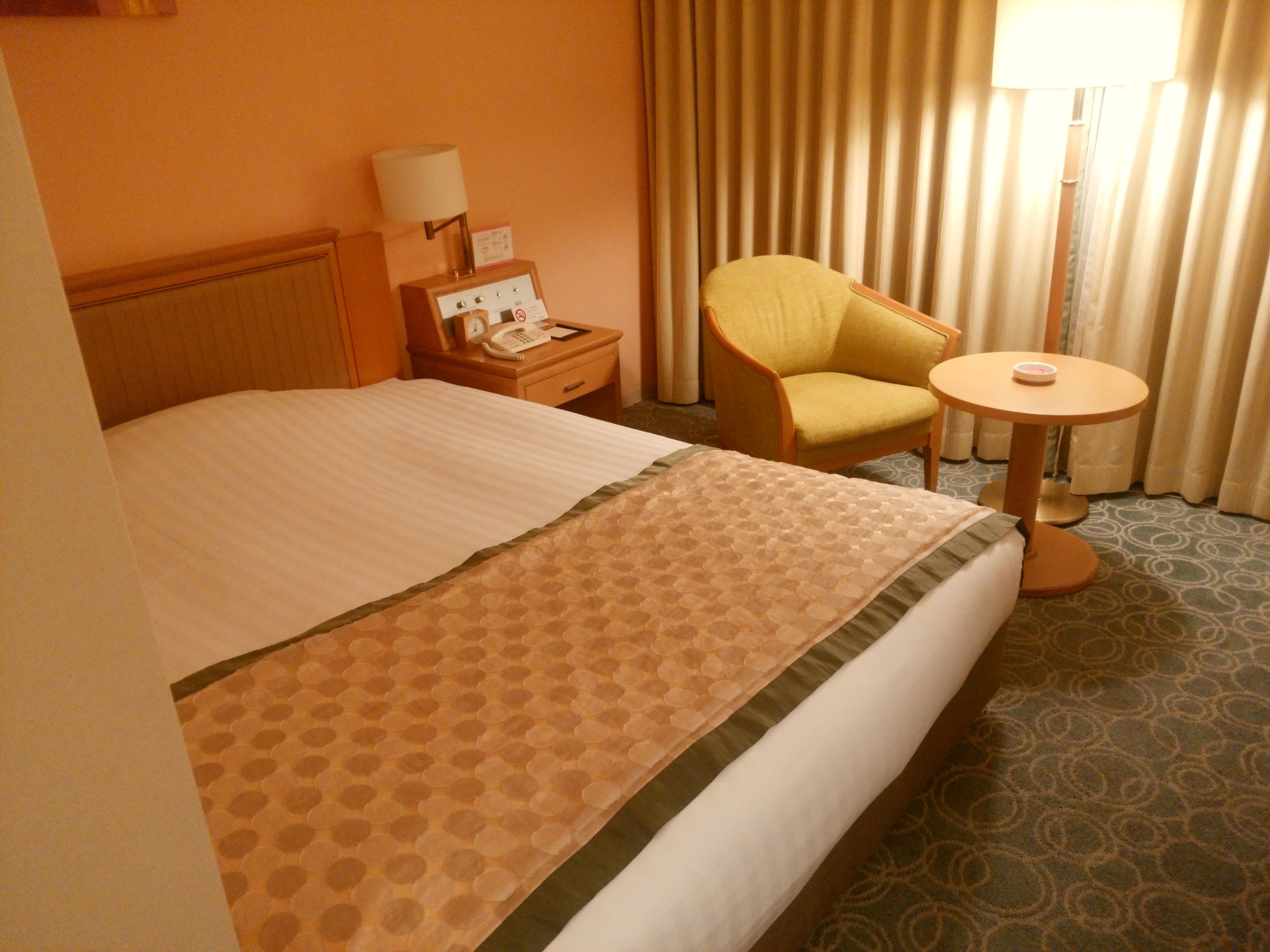 A hotel room with a bed, a chair, a bedside table with a lamp, a phone, and a small table with a cup.
