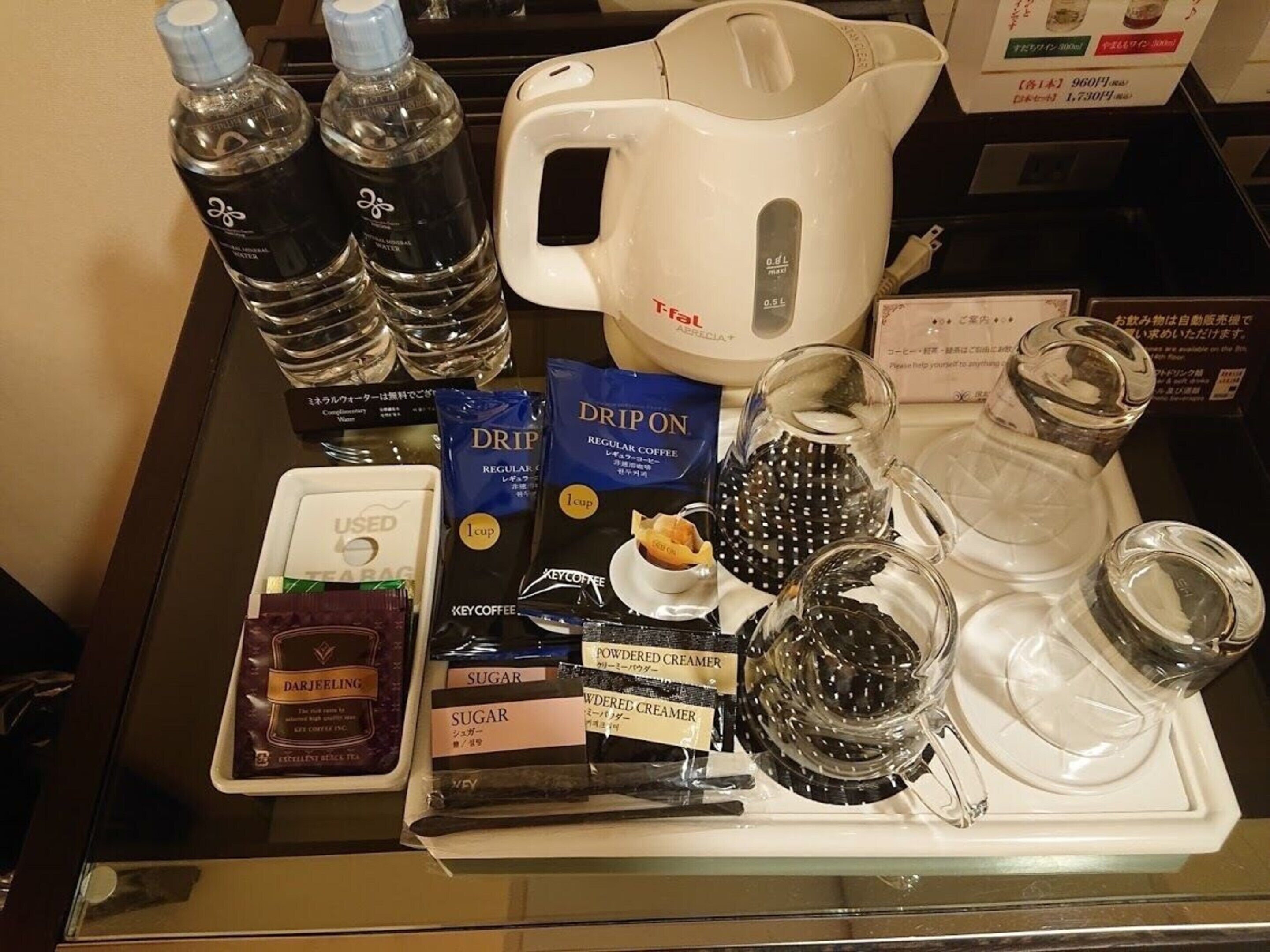 Una bandeja con utensilios para preparar café, incluyendo una cafetera de goteo, cápsulas de café, sobrecitos de azúcar, leche condensada y botellas de agua.