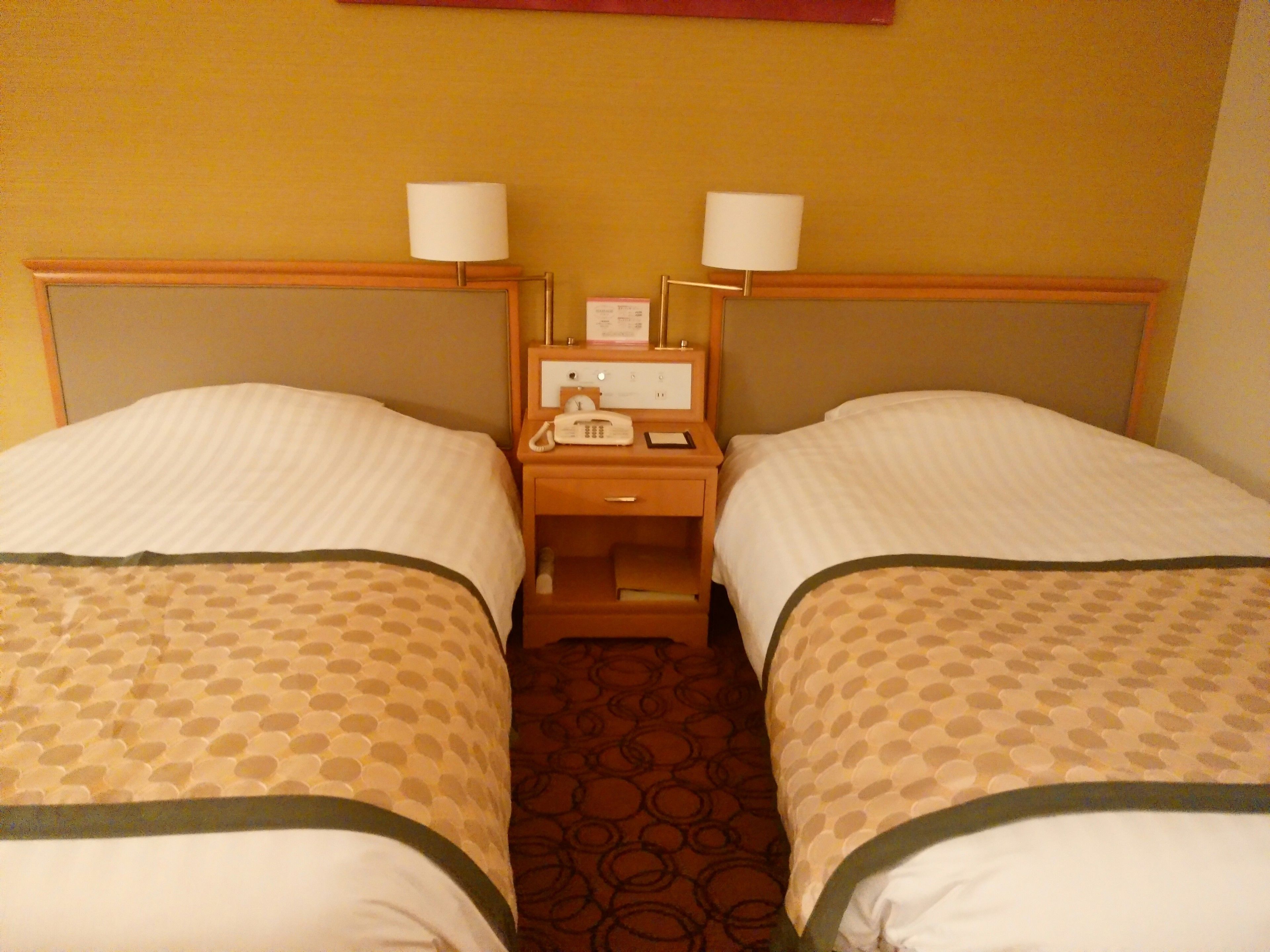 A hotel room with two single beds, each with a headboard and a bedside table with a lamp.