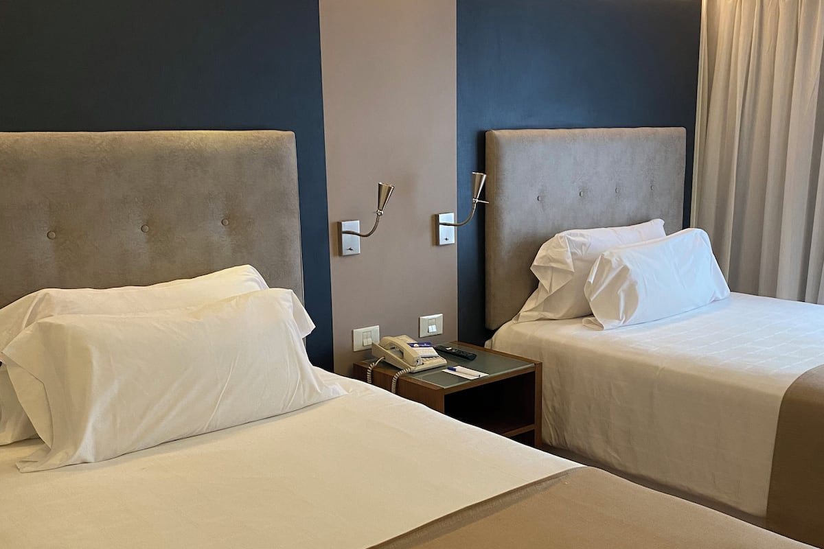 A hotel room with two beds, a bedside table with a phone and remote control, and a wall-mounted light fixture.