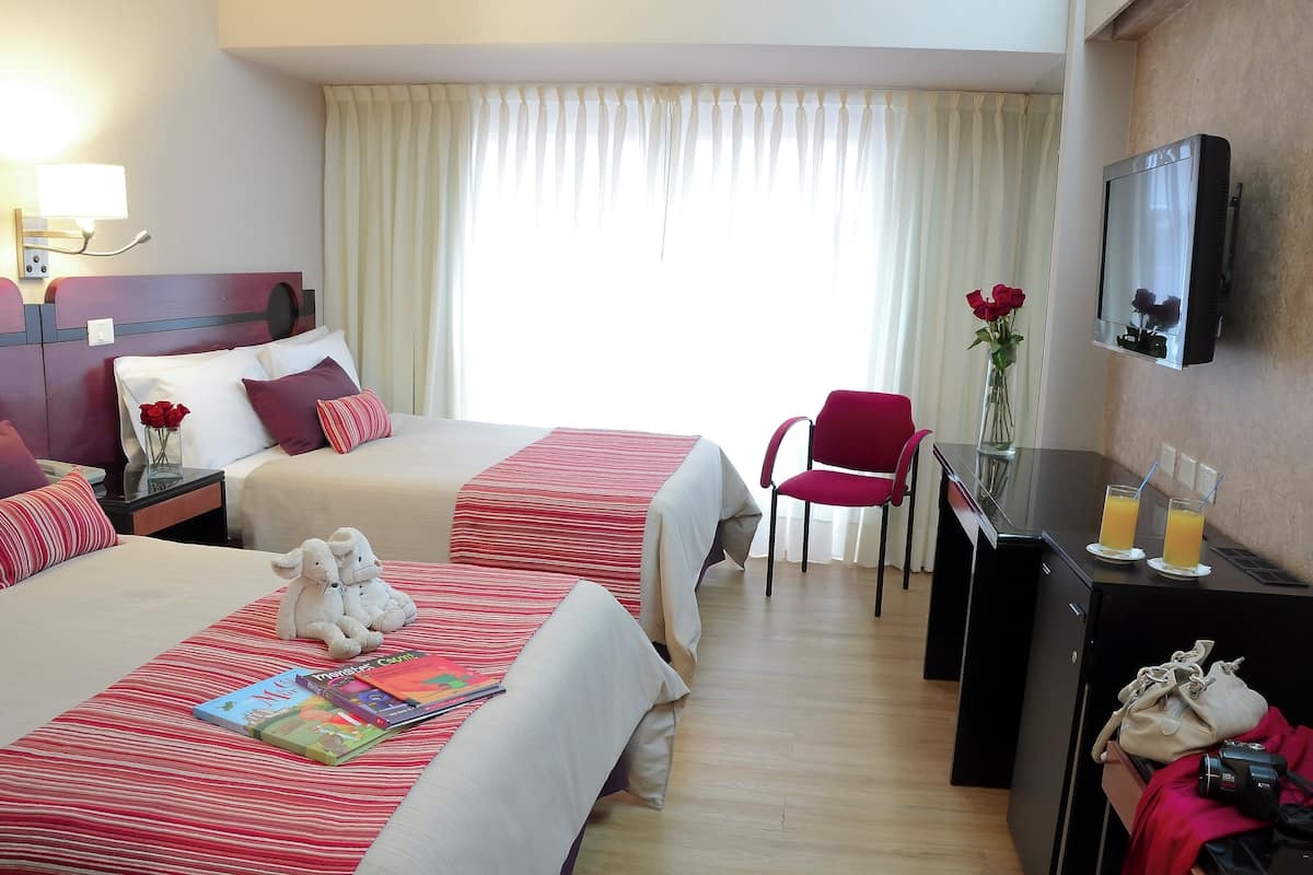 A hotel room with two beds, a desk with a mirror, a chair, and a small table with drinks and a vase of flowers.
