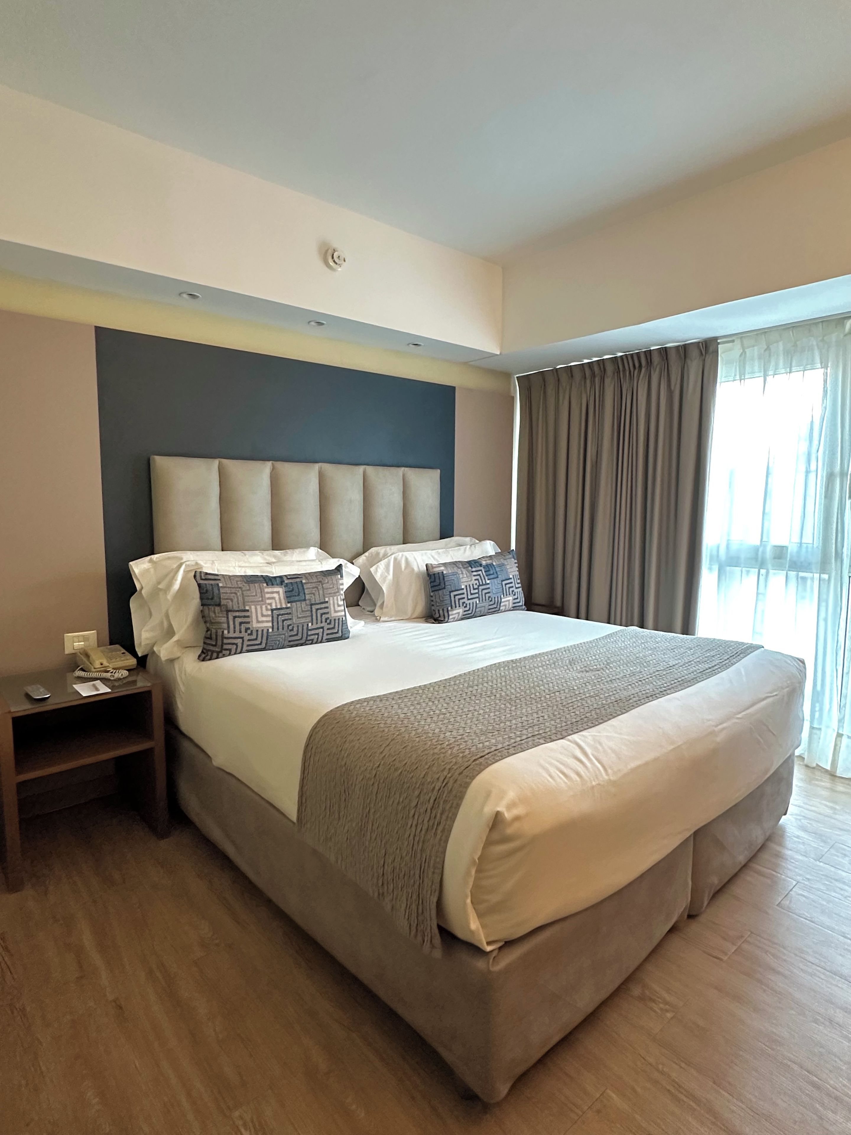 Habitación de hotel con una cama grande, una mesita de noche con un teléfono y documentos, y una ventana con cortinas.