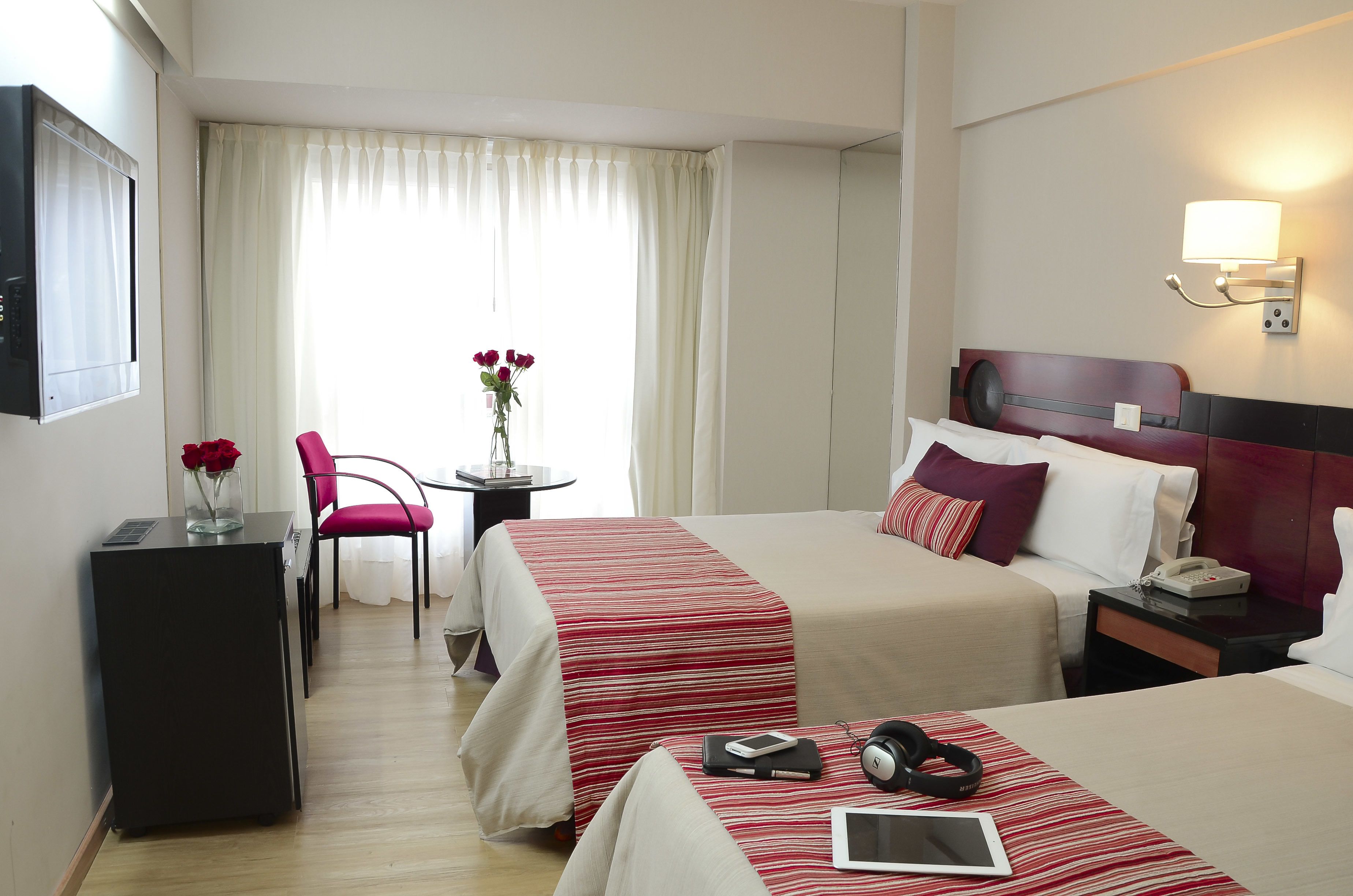 A hotel room with two beds, a desk, a chair, a small table with a vase of flowers, and a lamp on the wall.