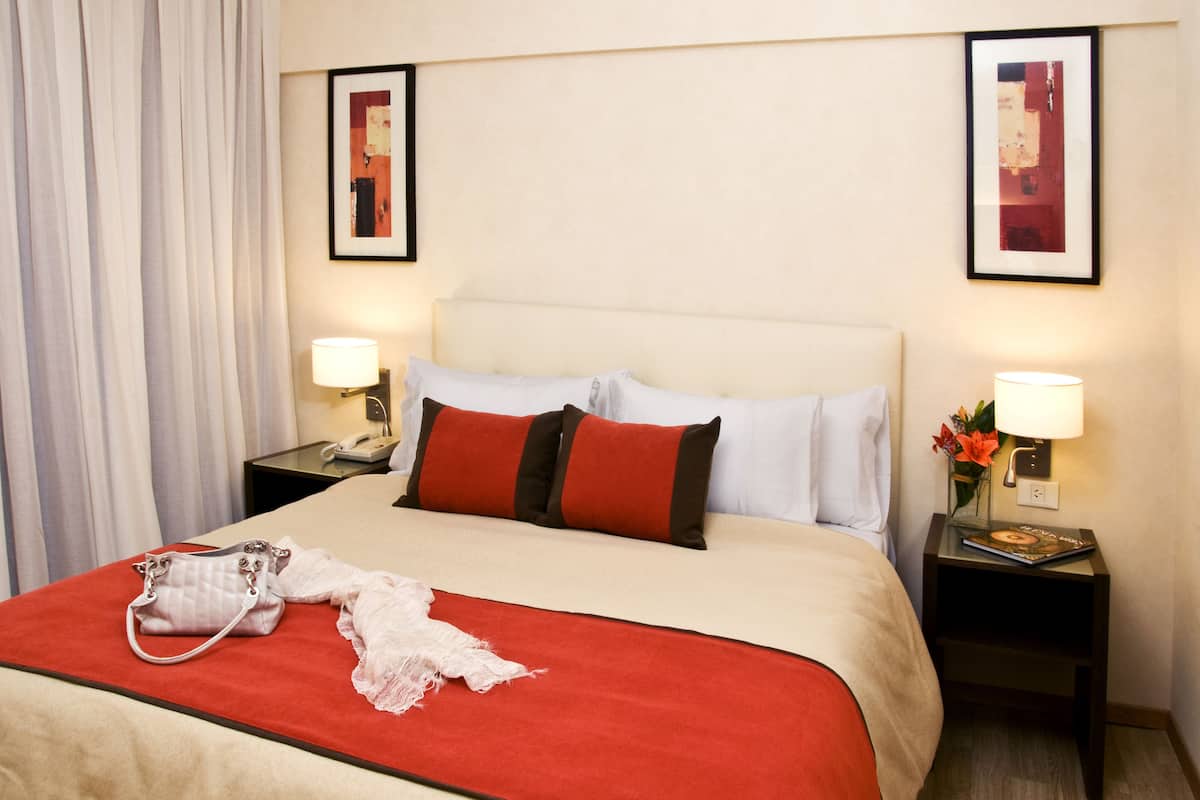 A hotel room with a bed, two bedside tables, a lamp on each table, a handbag on the bed, and a curtain on the window.
