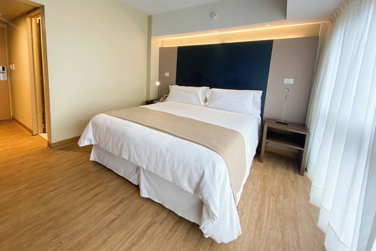 A hotel room with a large bed, bedside tables, a nightstand, a wall-mounted lamp, and a door leading to another room.