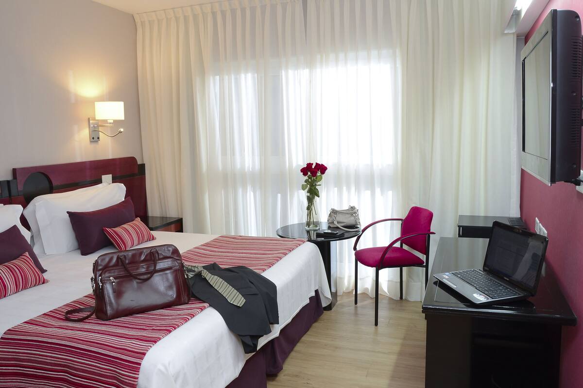 A hotel room with two beds, a desk with a laptop, a chair, a table with a vase and a handbag, and a window with curtains.