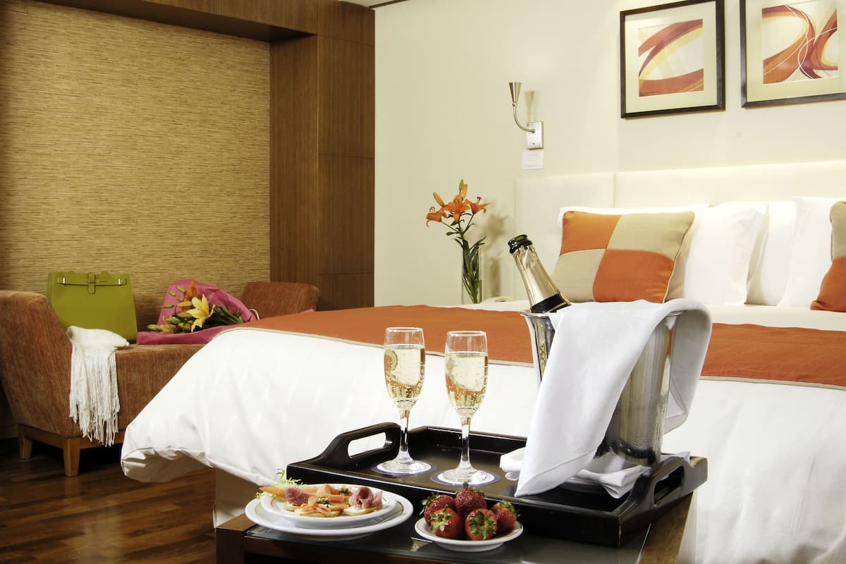 A hotel room with a bed, a dining table with a tray of food and drinks, and a vase with flowers.