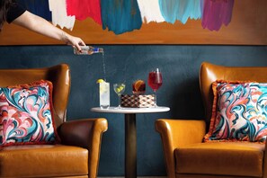 A table with two drinks and a bottle being poured, set between two leather chairs with colorful cushions. - Hyde & Seek, London Hyde Park, a Tribute Portfolio Hotel (London)