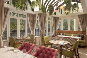 A dining area with round tables and chairs, large windows with curtains, and hanging plants. - Hyde & Seek, London Hyde Park, a Tribute Portfolio Hotel (London)