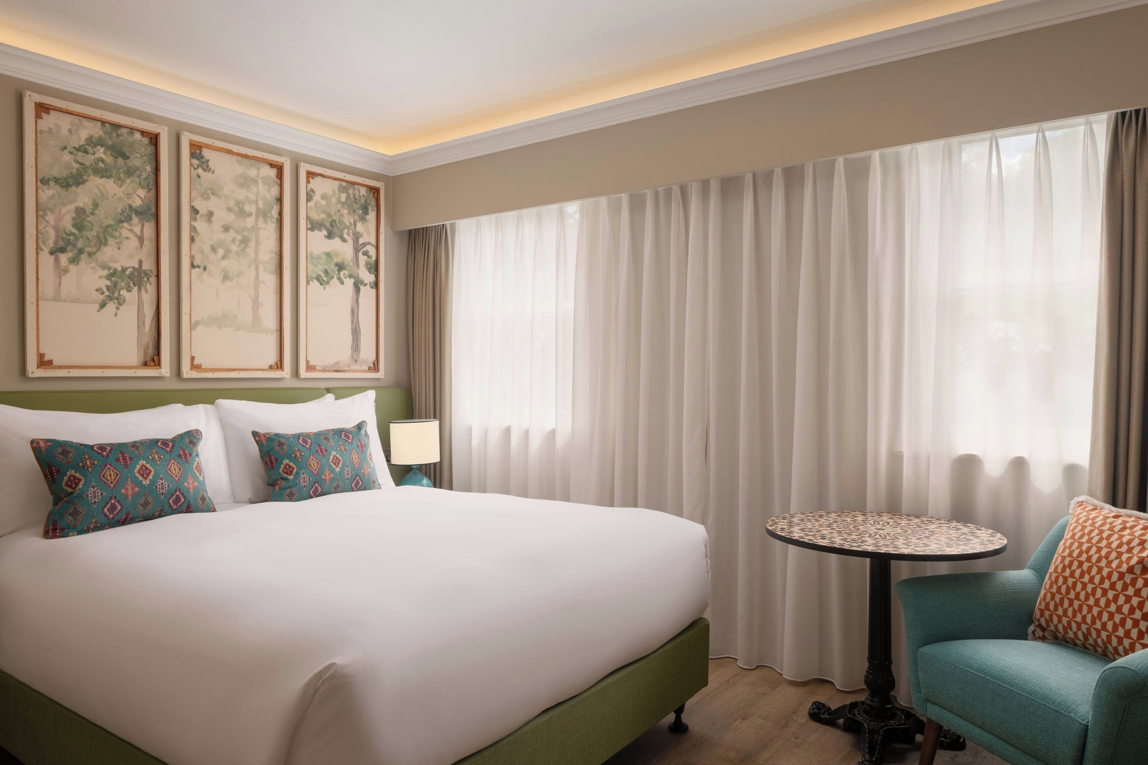 A hotel room with a large bed, a round table, and a chair. There are three framed paintings on the wall.