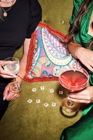 Una persona sostiene una copa de vino junto a un cojín colorido y un juego de Scrabble sobre un sofá verde.