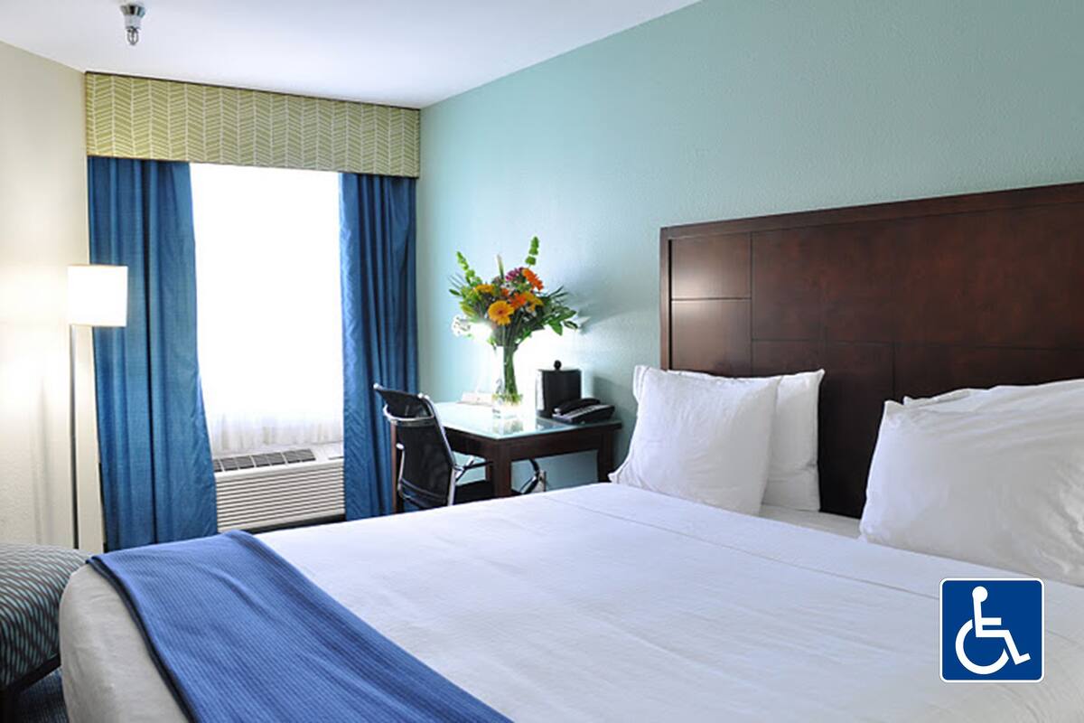 Una habitación de hotel con una cama grande, un escritorio con silla, un florero con flores y una ventana con cortinas.