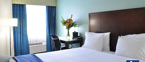 Ett hotellrum med en stor säng, ett skrivbord med en stol, en vas med blommor och ett fönster med gardiner.