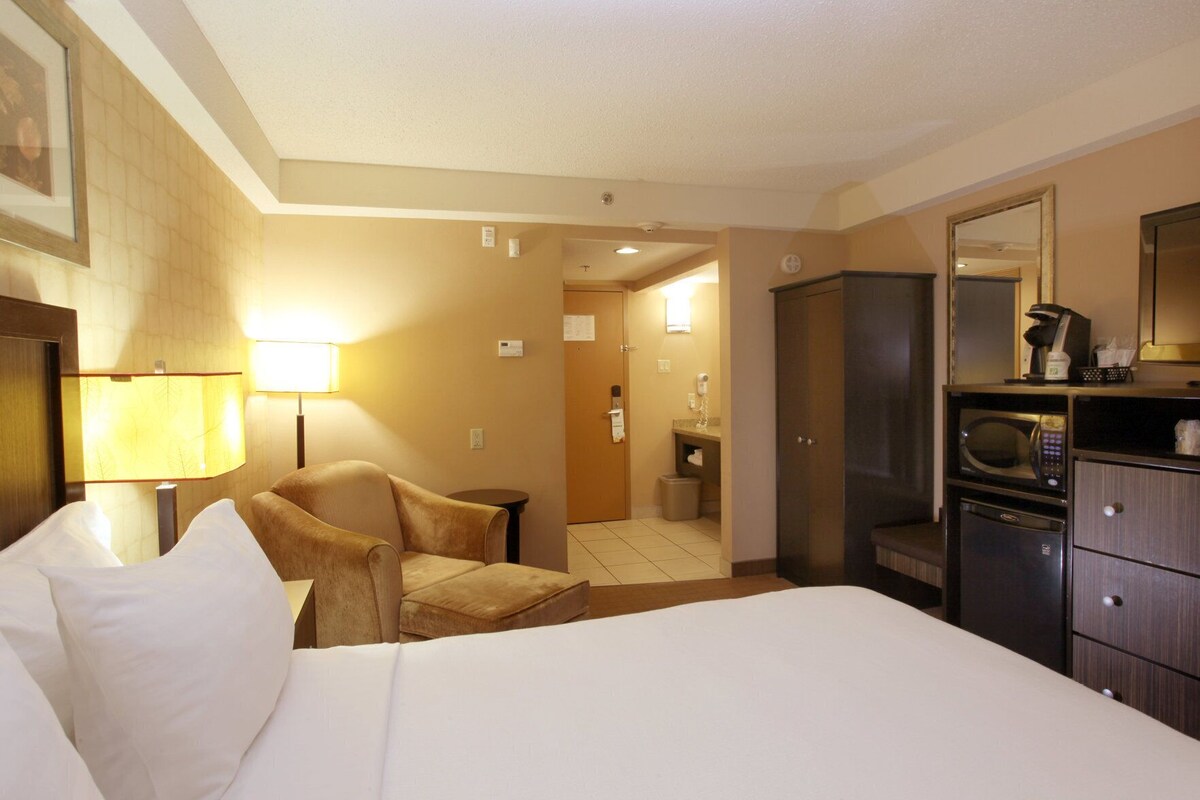 A hotel room with a bed, a chair, a desk, a microwave, a refrigerator, and a mirror.