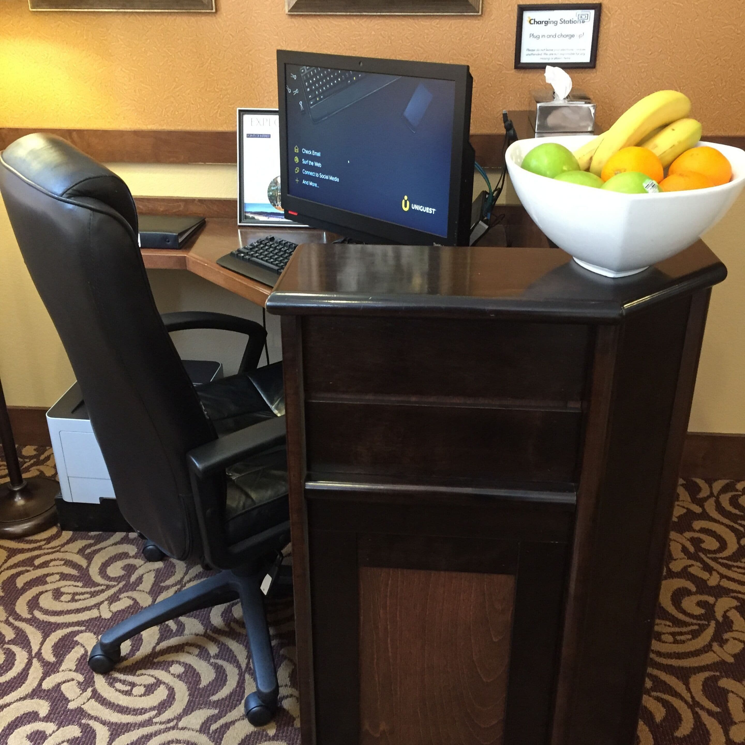 A hotel reception desk with a computer, a keyboard, a chair, a printer, and a bowl of fruit.