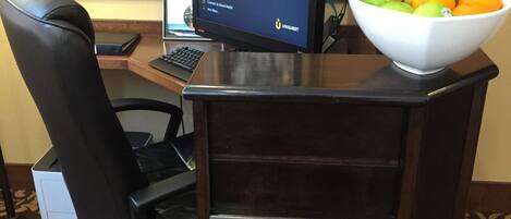 A hotel reception desk with a computer, a keyboard, a chair, a printer, and a bowl of fruit.