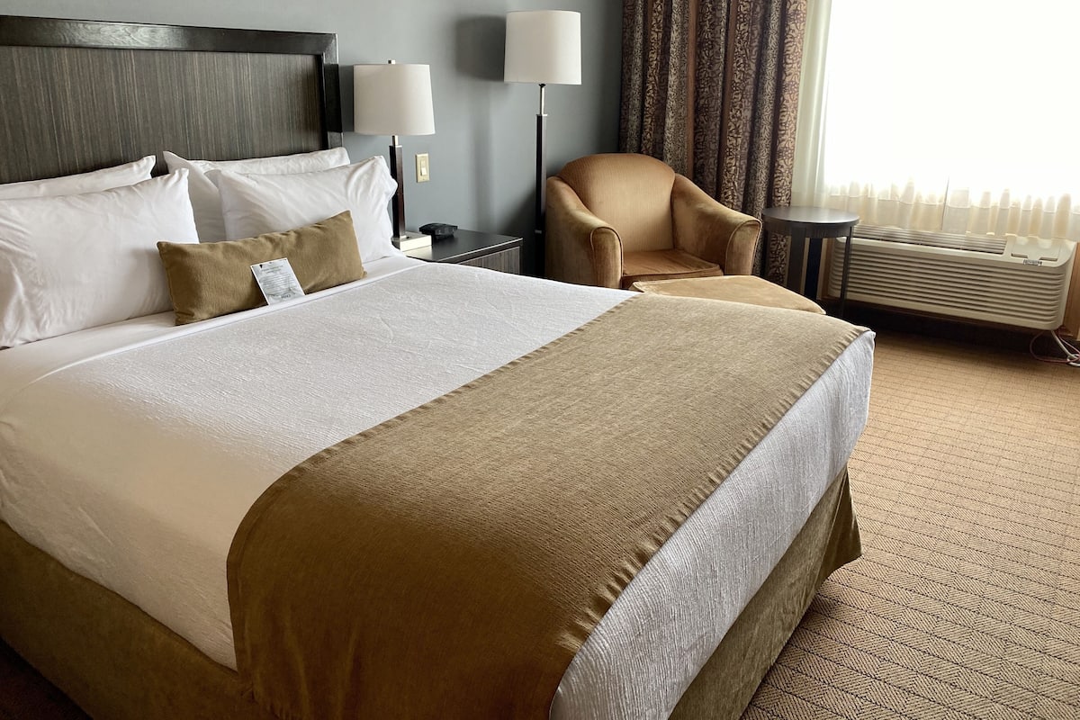 A hotel room with a large bed, two bedside lamps, a chair, an air conditioning unit, and a window with curtains.