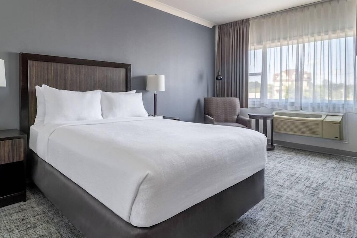 A hotel room with a large bed, two bedside tables with lamps, a chair, a small table, and a window with curtains.