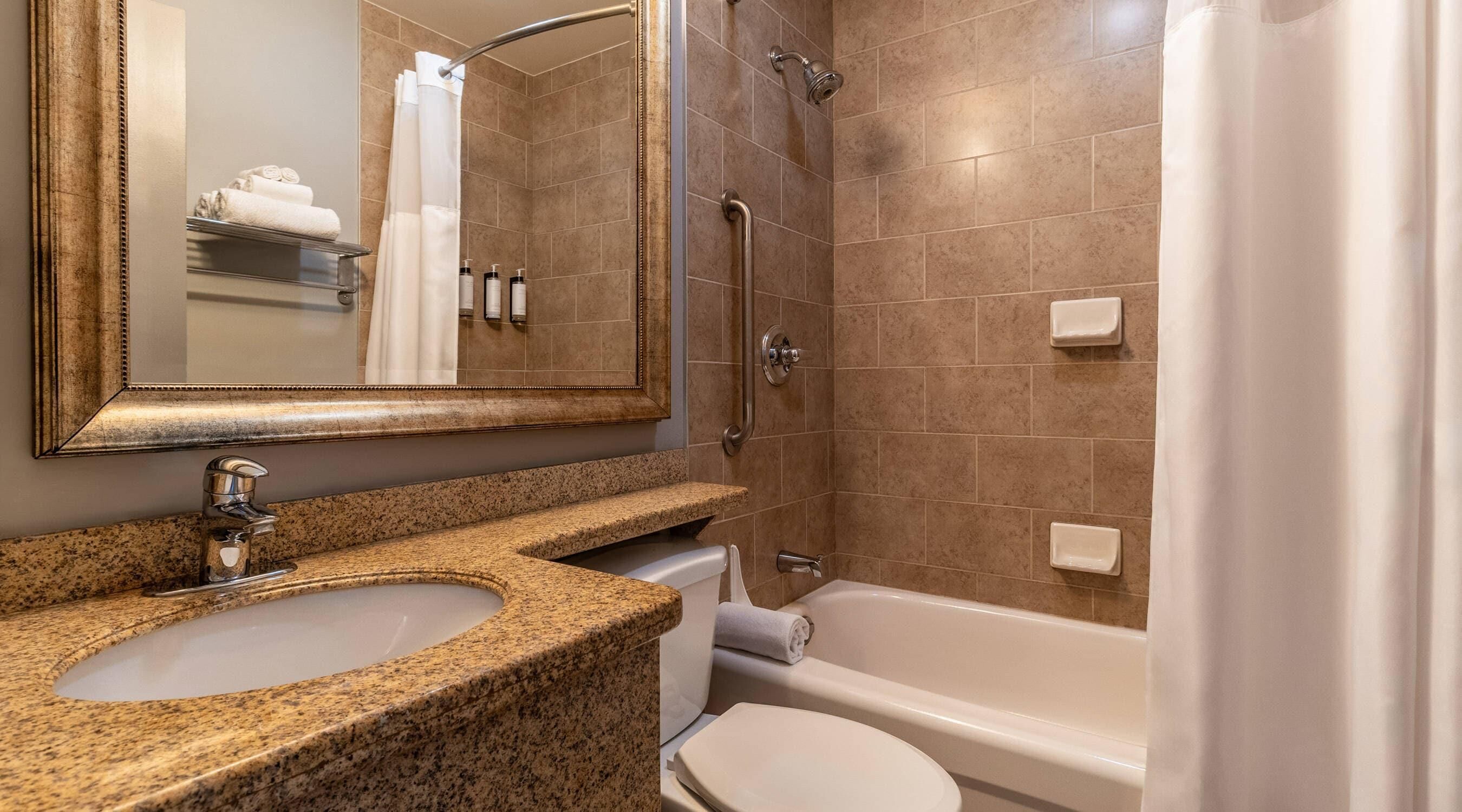 A bathroom with a granite countertop sink, a toilet, a bathtub, and a shower with a grab bar.