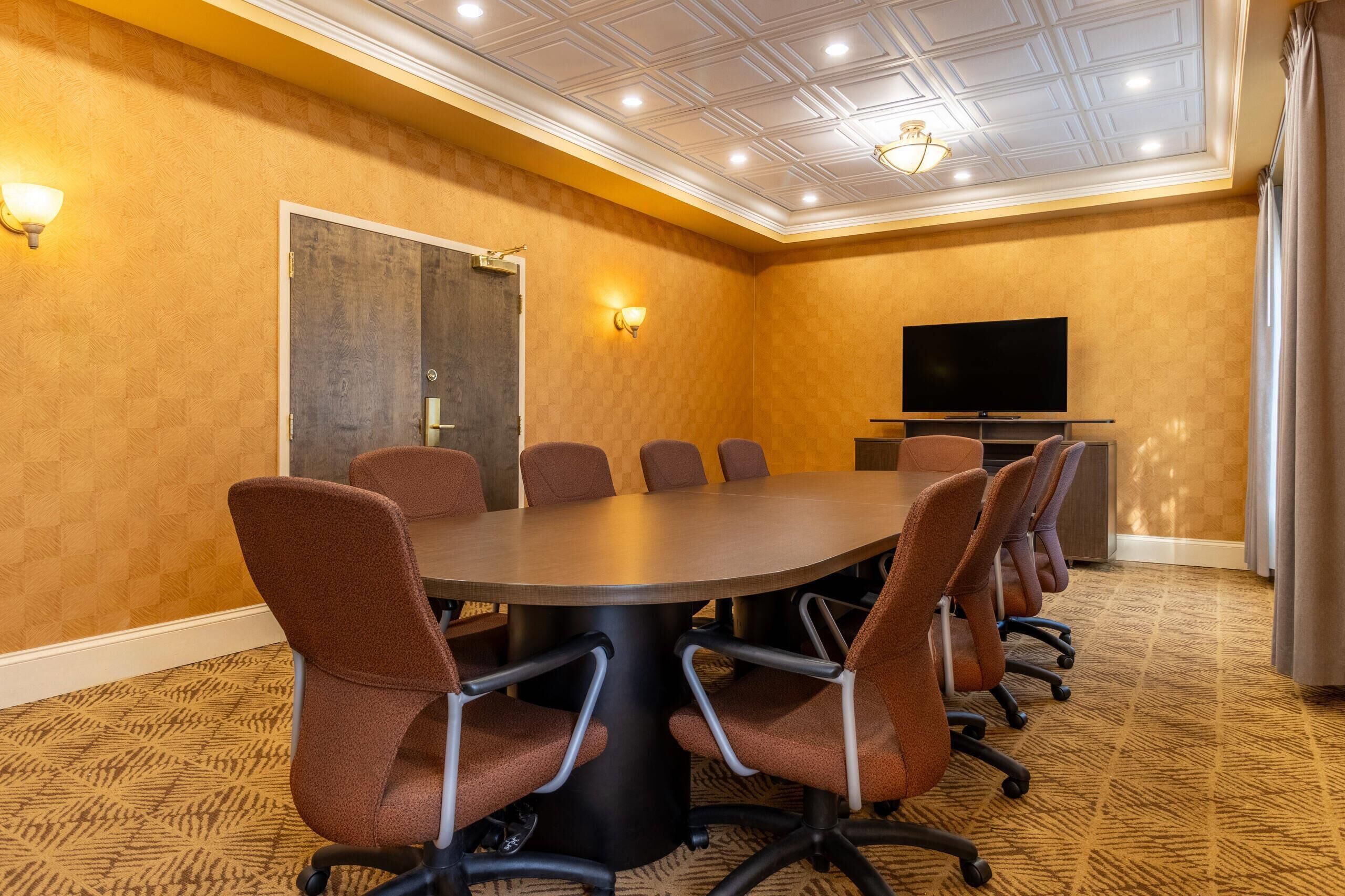 A conference room with a large oval table, six brown chairs, a flat-screen TV, and a door with a light fixture.