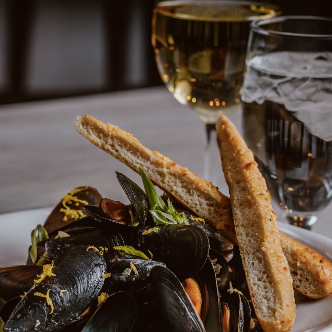 A plate of mussels with lemon zest, accompanied by slices of bread and a glass of white wine.