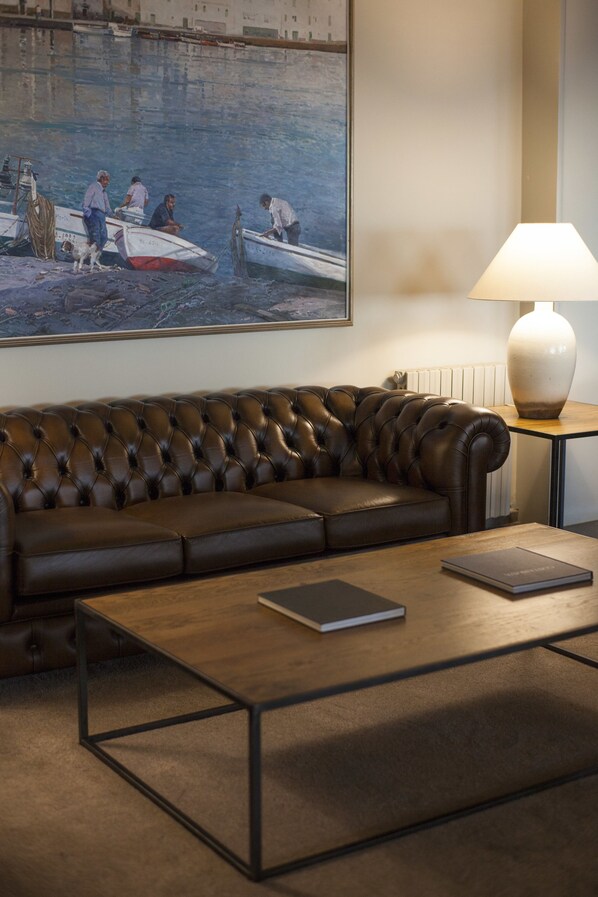 A brown leather sofa, a wooden coffee table, and a painting of boats on a wall. - Hotel Santa Marta (Lloret de Mar)