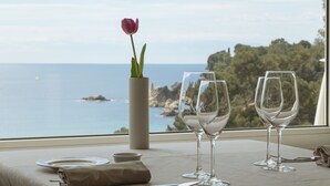 Un tavolo apparecchiato con bicchieri da vino, un vaso con un singolo fiore e vista sul mare e sulla costa rocciosa attraverso una finestra.