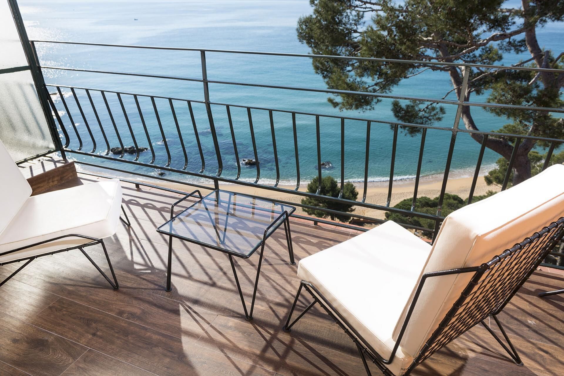 A balcony with a view of the sea, wooden flooring, and two white chairs.