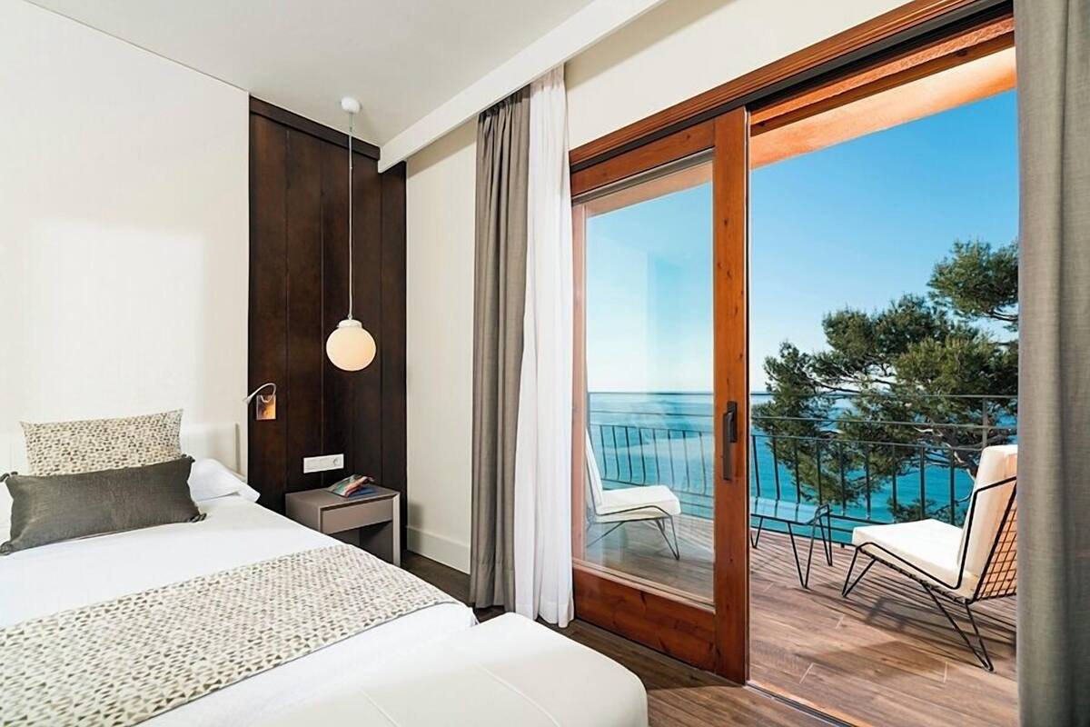 Hotelkamer met een bed, nachtkastje, lamp en een balkon met uitzicht op zee.