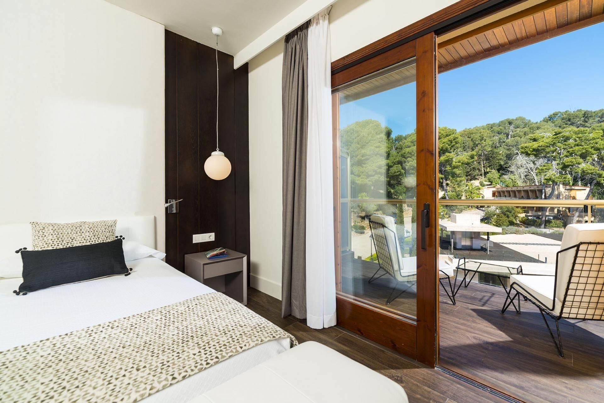 Una habitación de hotel moderna con una cama grande, una mesita de noche, una lámpara de lectura y un ventanal corredero que da a un balcón con mobiliario exterior.