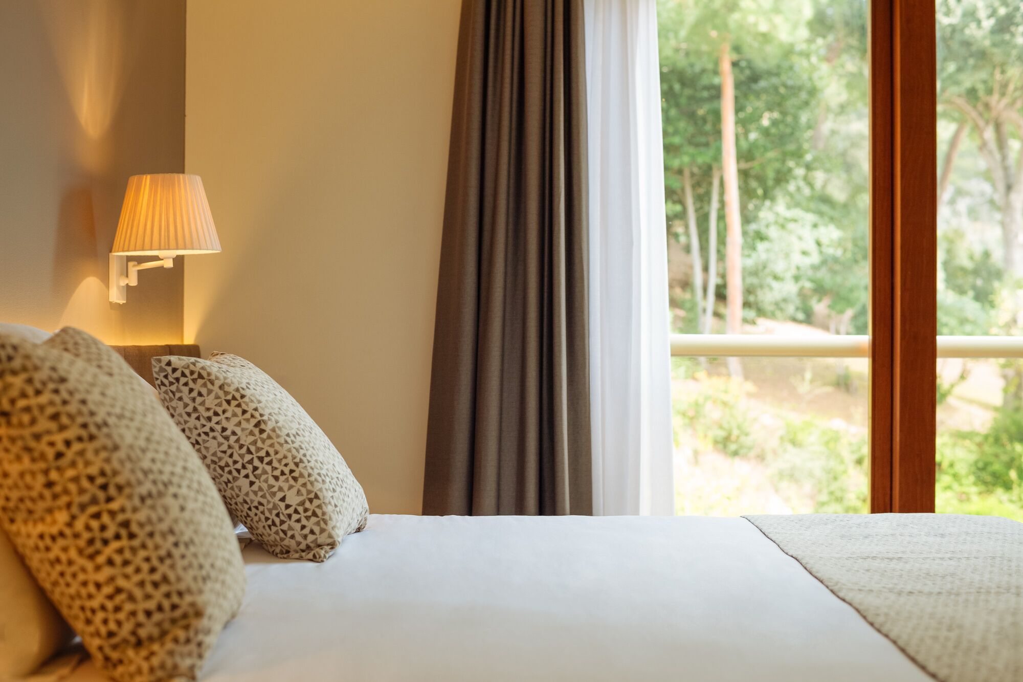A hotel room with a bed, a lamp, and a view of greenery through the window.
