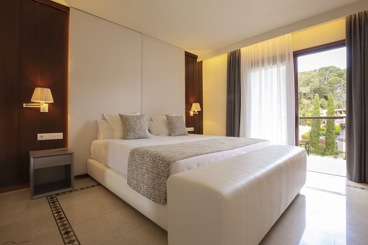 A modern hotel room with a large bed, two bedside lamps, a nightstand, and a view of the outdoors.