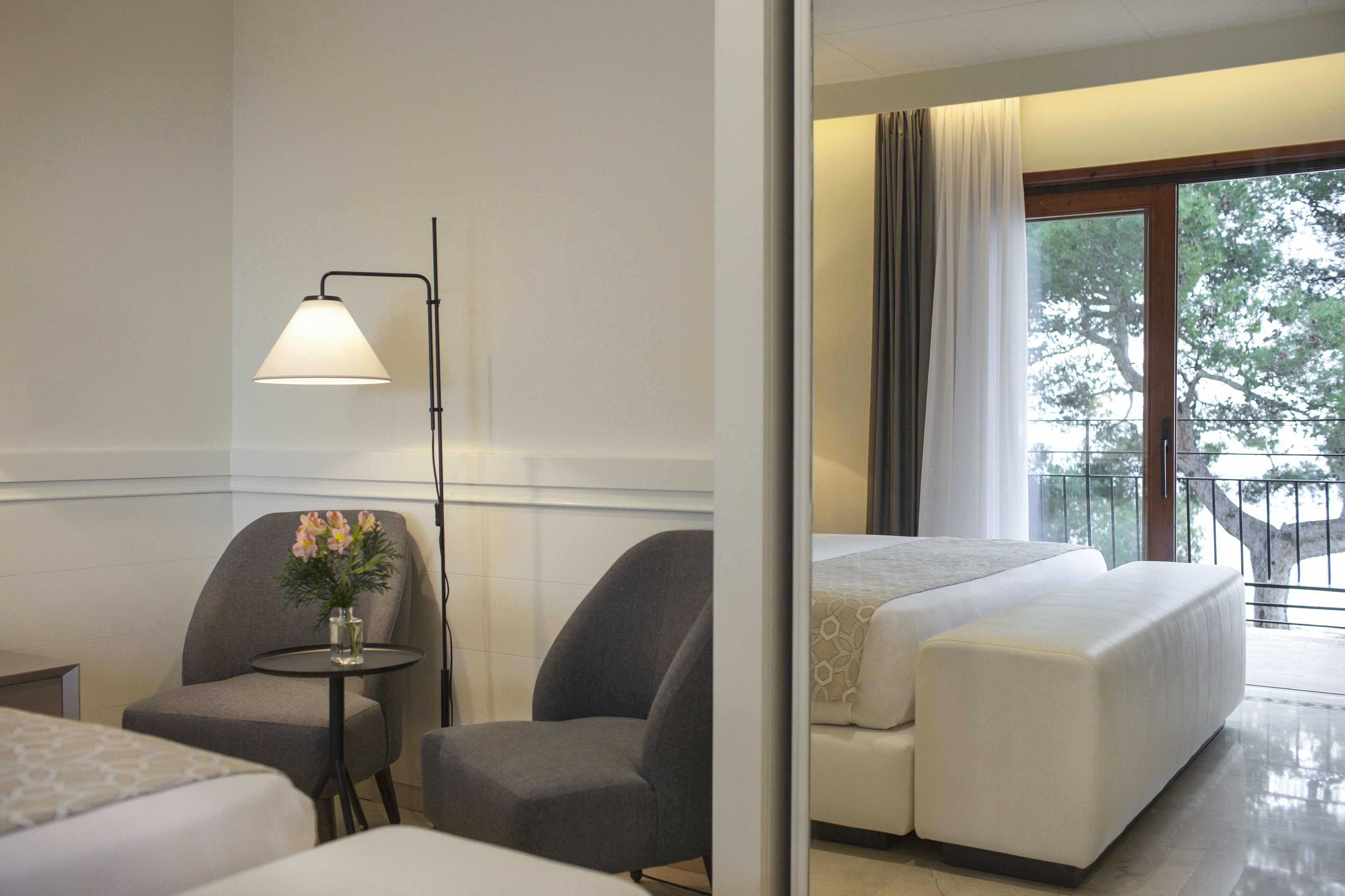 Ett hotellrum med en säng, två fåtöljer, ett litet bord med en vas med blommor och en golvlampa.