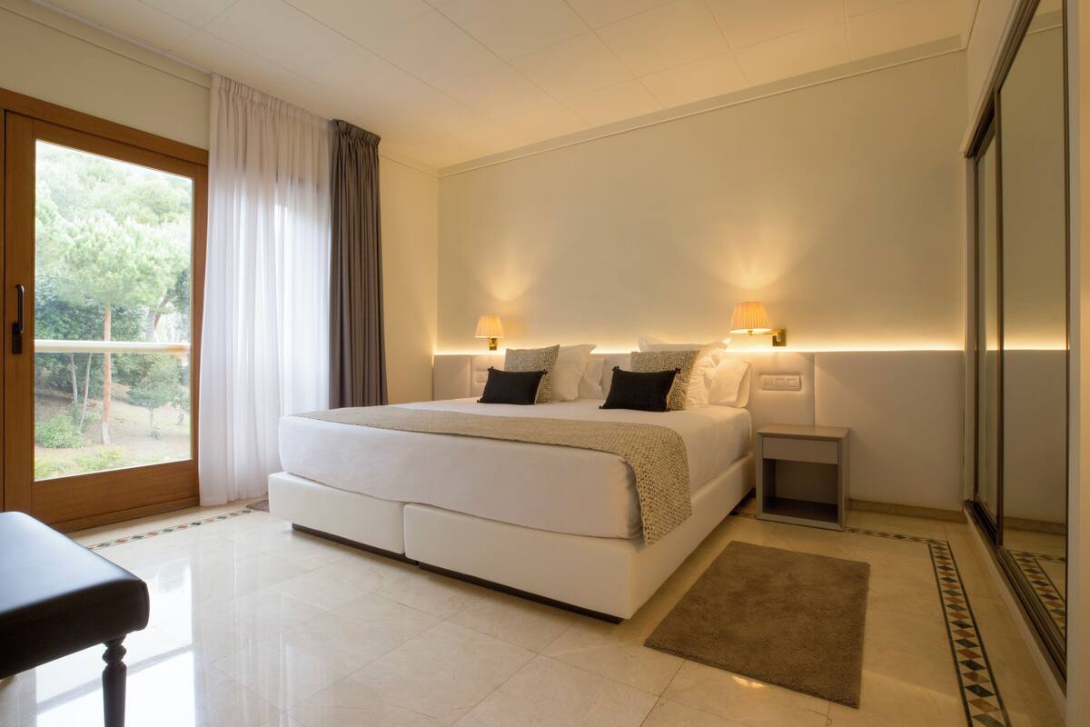 A modern bedroom with a large bed, bedside lamps, a small nightstand, and a view of greenery outside.