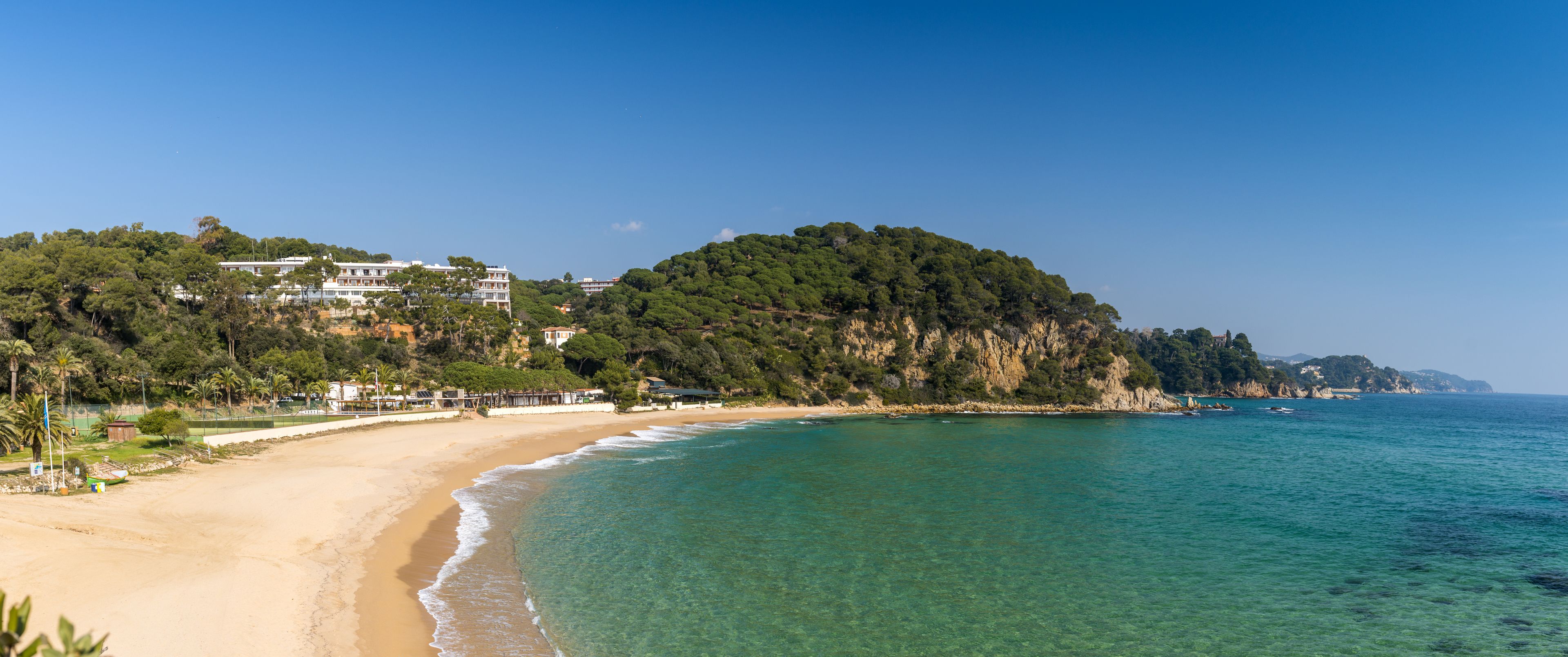 A coastal beach with clear turquoise waters, a sandy shore, and a hillside with buildings and greenery.