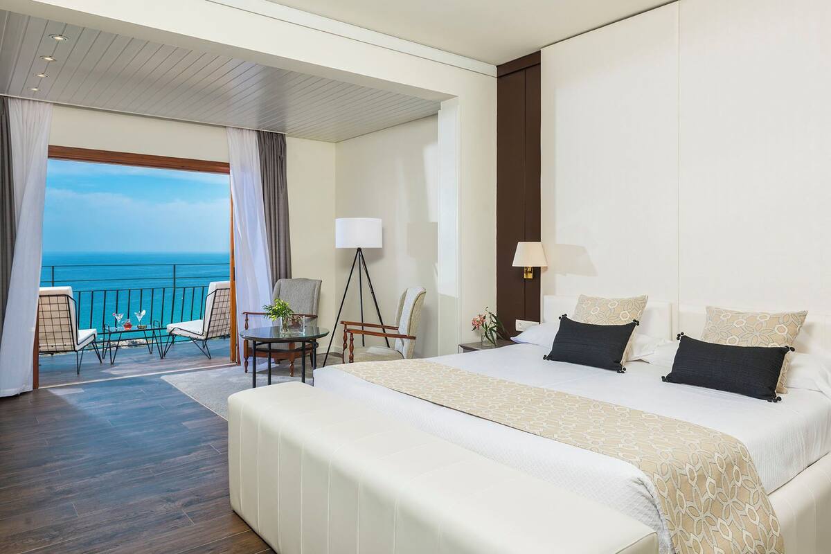 A hotel room with a large bed, a balcony with a view of the sea, a sitting area with chairs, and a small table.