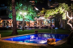 A pool area with a clear pool, surrounded by grass and trees, and a building with outdoor seating in the background. - Manquehue Las Condes Hotel (Santiago)