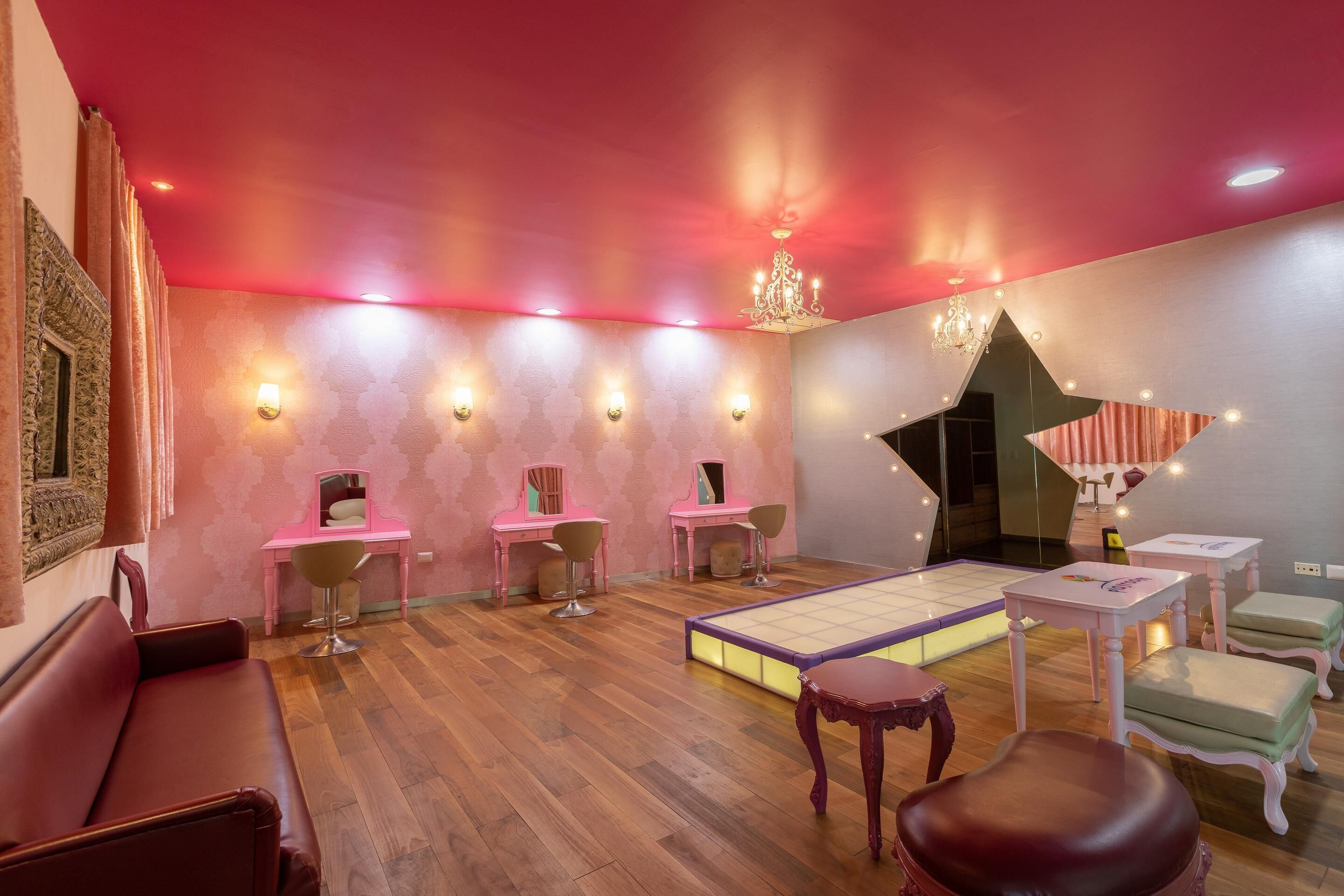 A room with a wooden floor, pink walls, and a red ceiling. There are three pink desks with chairs, a large mirror, and a chandelier.
