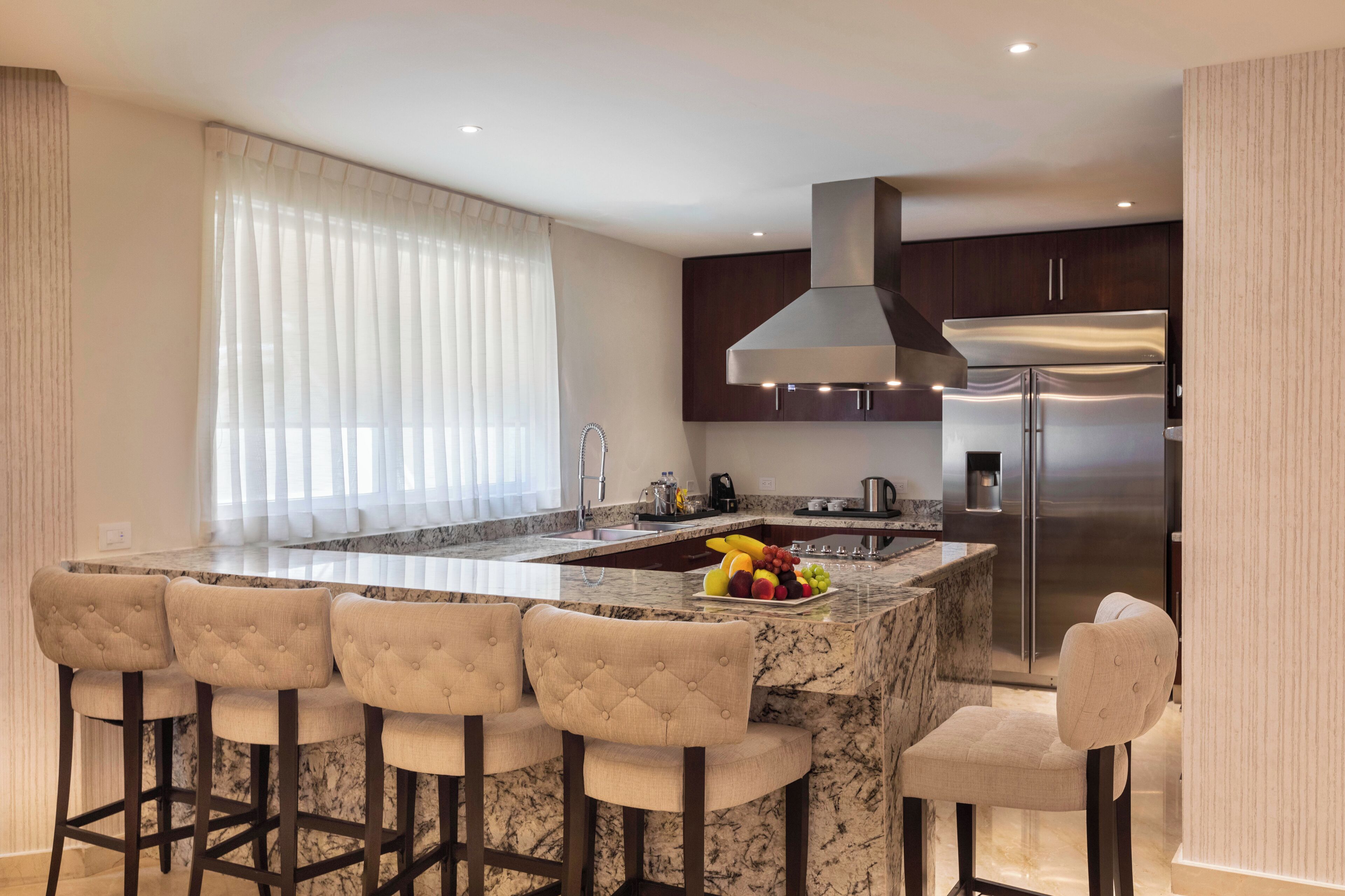A modern kitchen with a central island, bar stools, stainless steel appliances, and a large window with sheer curtains.