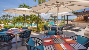 Un patio al aire libre con mesas y sillas, sombrillas y vistas a una piscina y una playa.