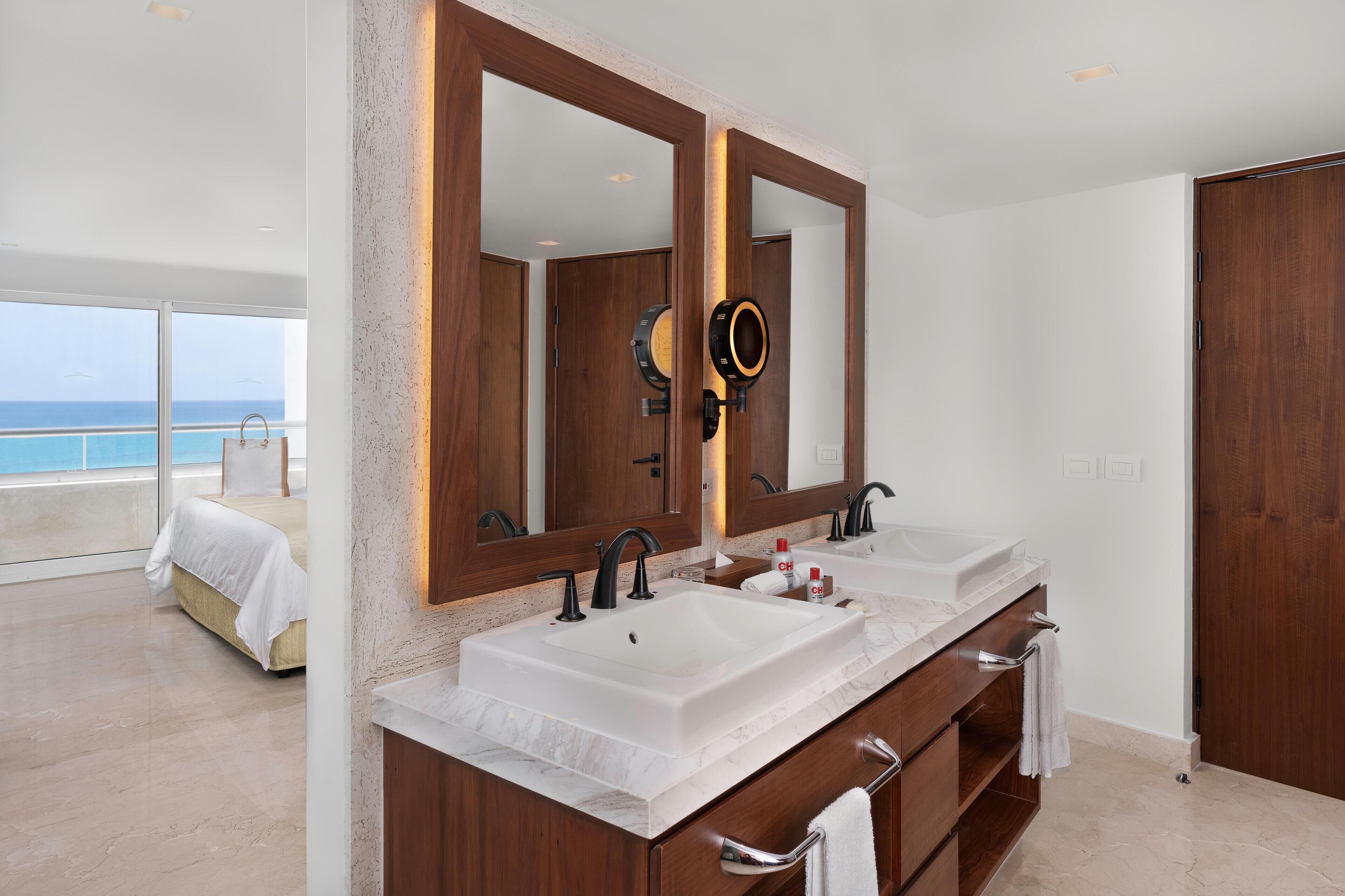 A modern bathroom with dual sinks, large mirrors, and a view of the ocean.